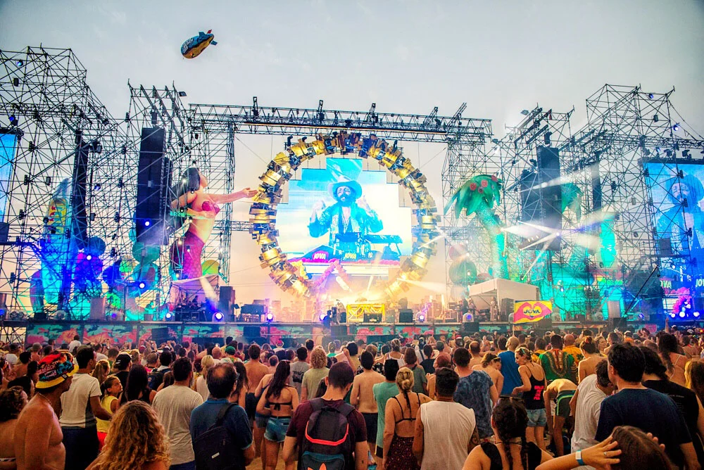 Palco del Jova Beach di giorno: anello di luci attorno a uno schermo, enormi figure di sirena e palma sulle torri di tubi – Jova Beach Party 2019, Italia – scenografia per concerti