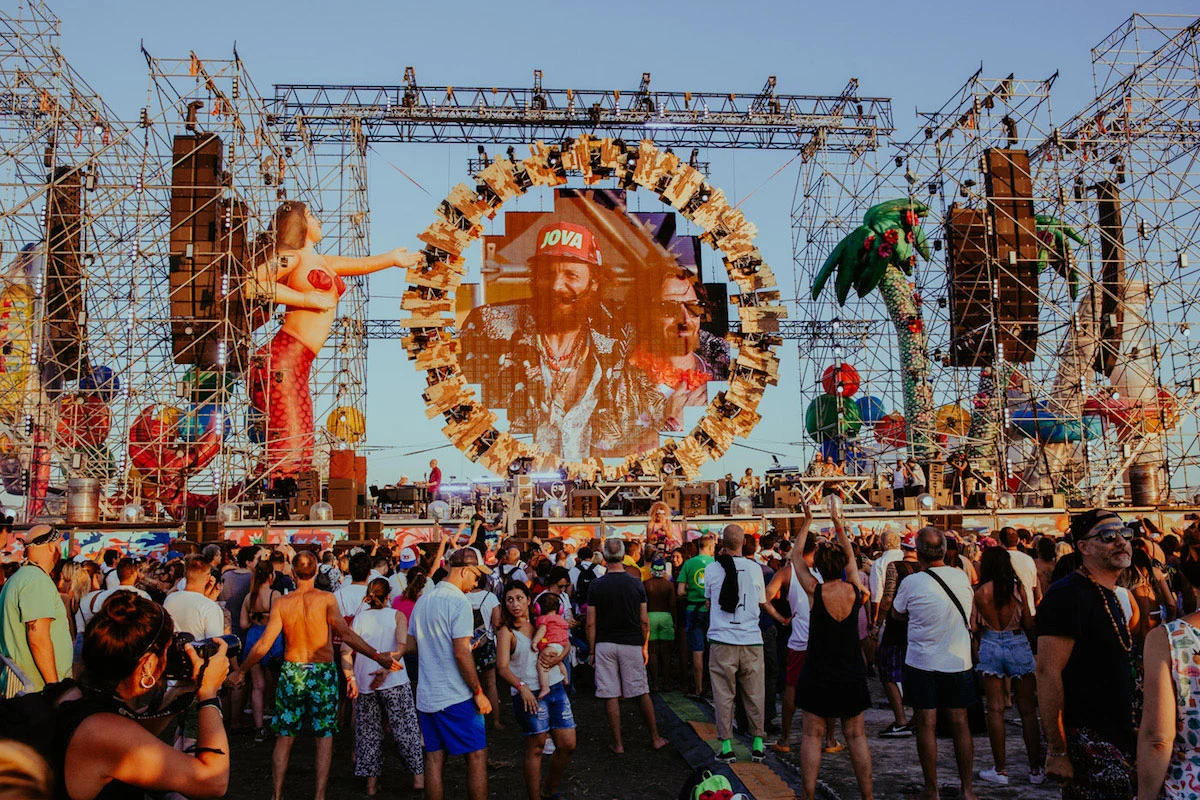 Pubblico al sole del pomeriggio davanti al palco, con una figura di sirena e palme sulle torri di tubi – Jova Beach Party 2019, Italia – scenografia per concerti