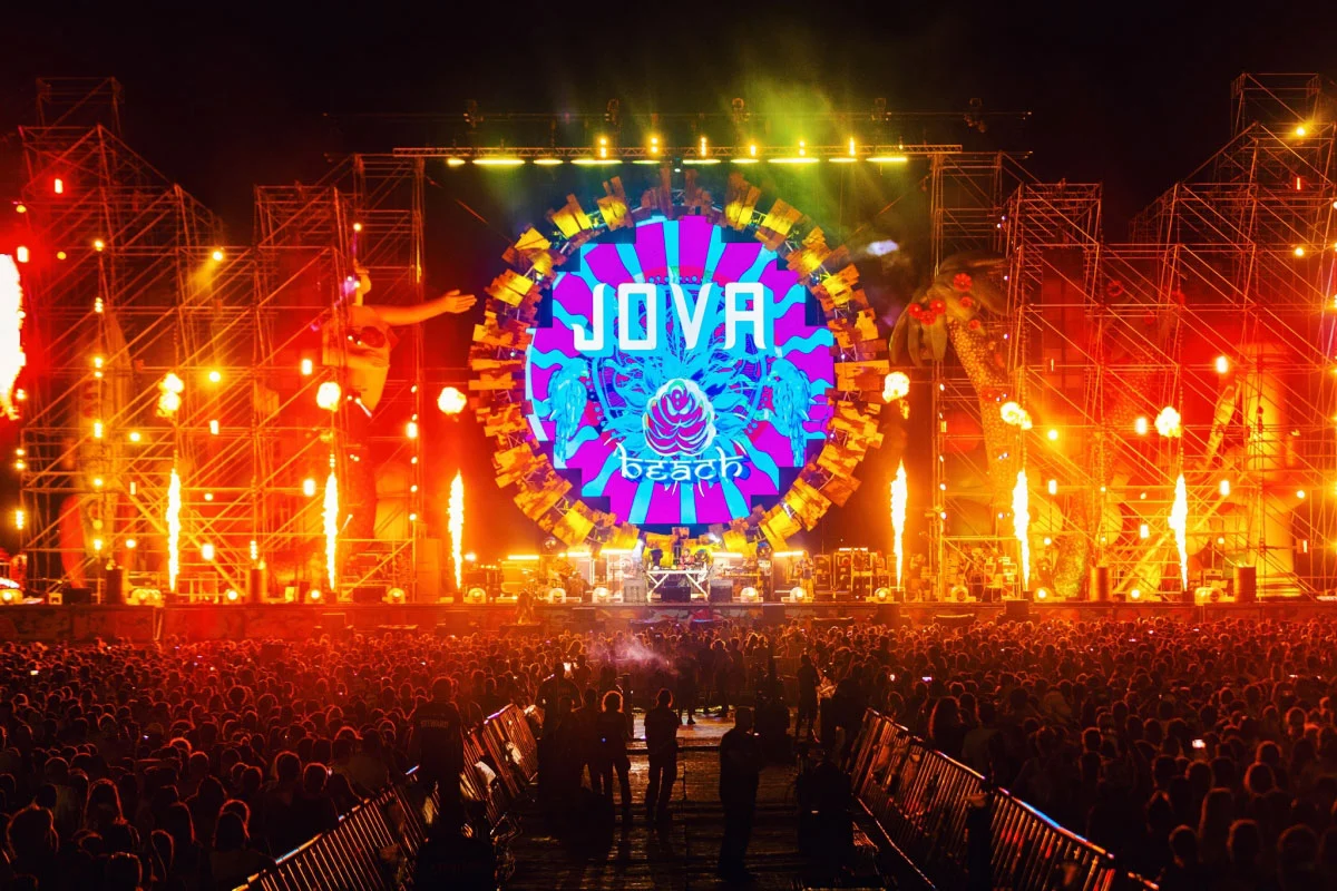 Vista notturna del palco del Jova Beach: schermo circolare con il logo tra torri di tubi, fiamme e un grande pubblico – Jova Beach Party 2019, Italia – scenografia per concerti