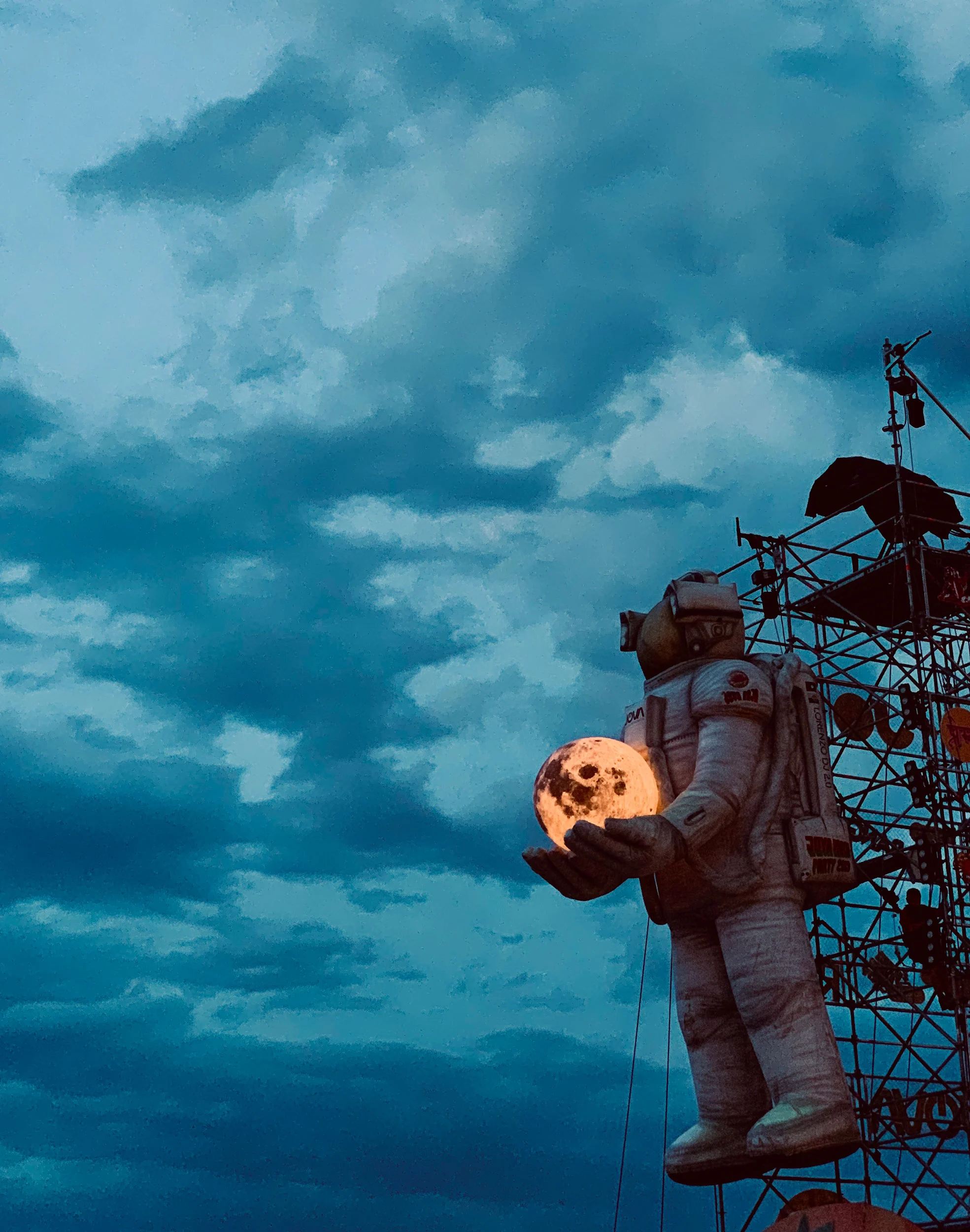 Astronauta gonfiabile che regge una luna luminosa sulla torre di tubi contro un cielo serale nuvoloso – Jova Beach Party 2019, Italia – scenografia per concerti