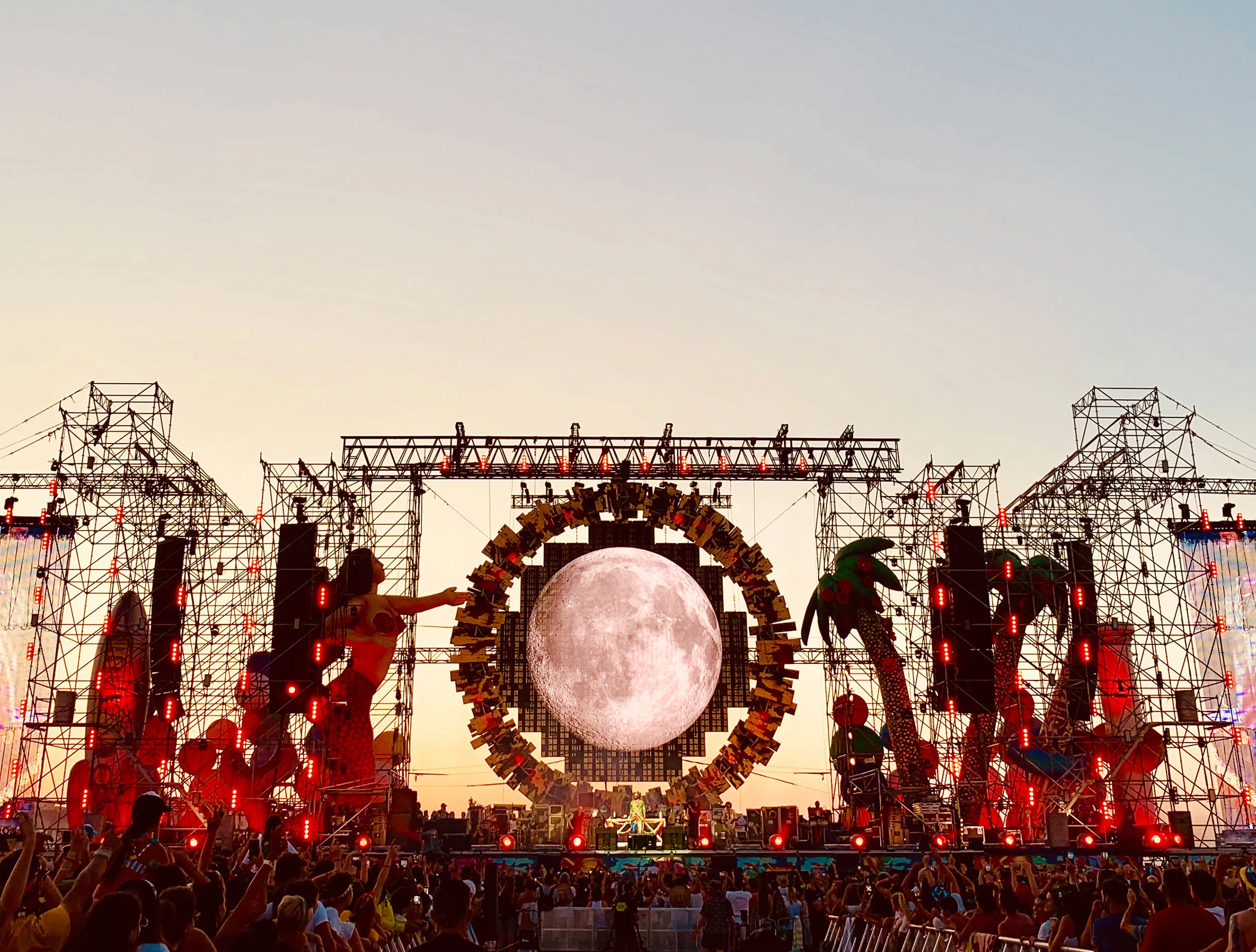 Palco al crepuscolo con l'immagine di una luna piena dentro l'anello, figure di sirena e palma su torri in luce rossa – Jova Beach Party 2019, Italia – scenografia per concerti