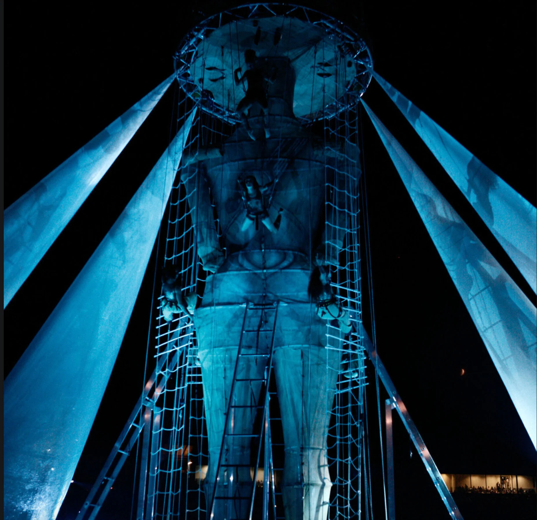 Vista dal basso della figura gigante nella sua struttura di reti e scale, tra vele di tessuto illuminate di blu – Cerimonia di apertura dei Giochi del Mediterraneo Pescara 2009, Italia, 2019 – progettazione di eventi