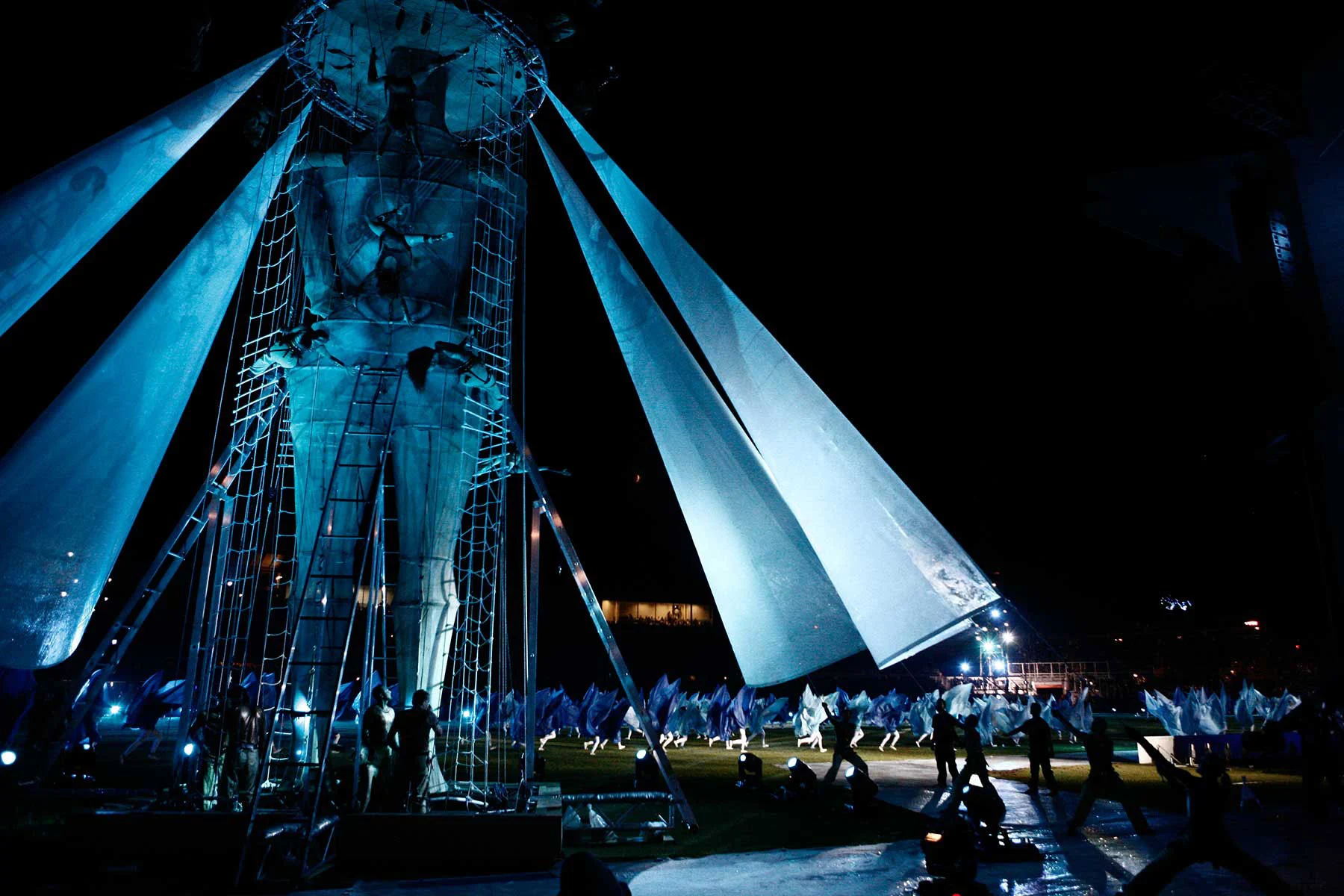 Alta figura simile a una statua in una struttura di scale e reti con ali di tessuto simili a vele, illuminata di blu – Cerimonia di apertura dei Giochi del Mediterraneo Pescara 2009, Italia, 2019 – progettazione di eventi