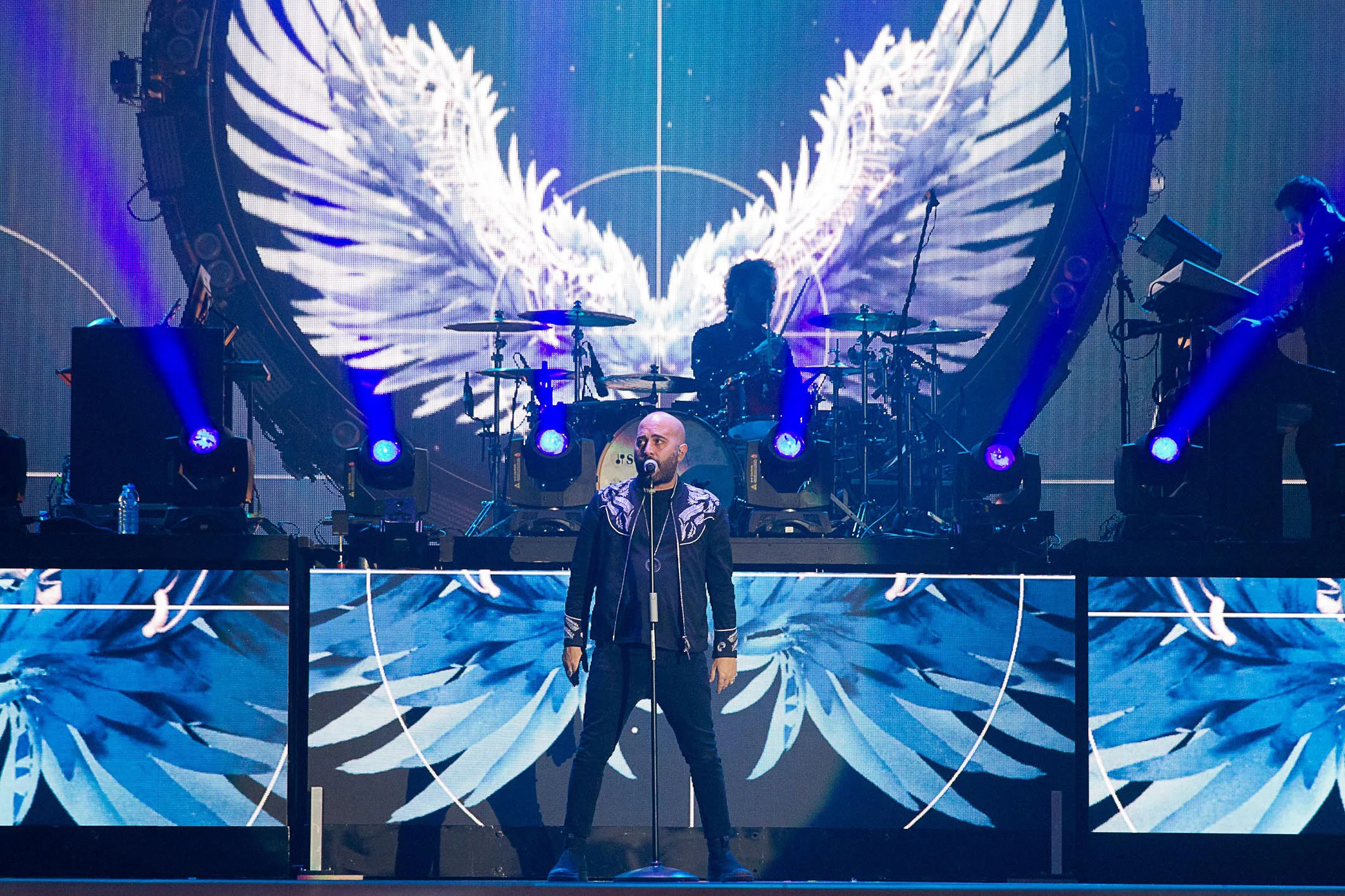 Il cantante dei Negramaro sul fronte del palco, ali di piume bianche proiettate sugli schermi dietro la batteria – Negramaro Amore che Torni Tour 2018, Italia – scenografia per concerti