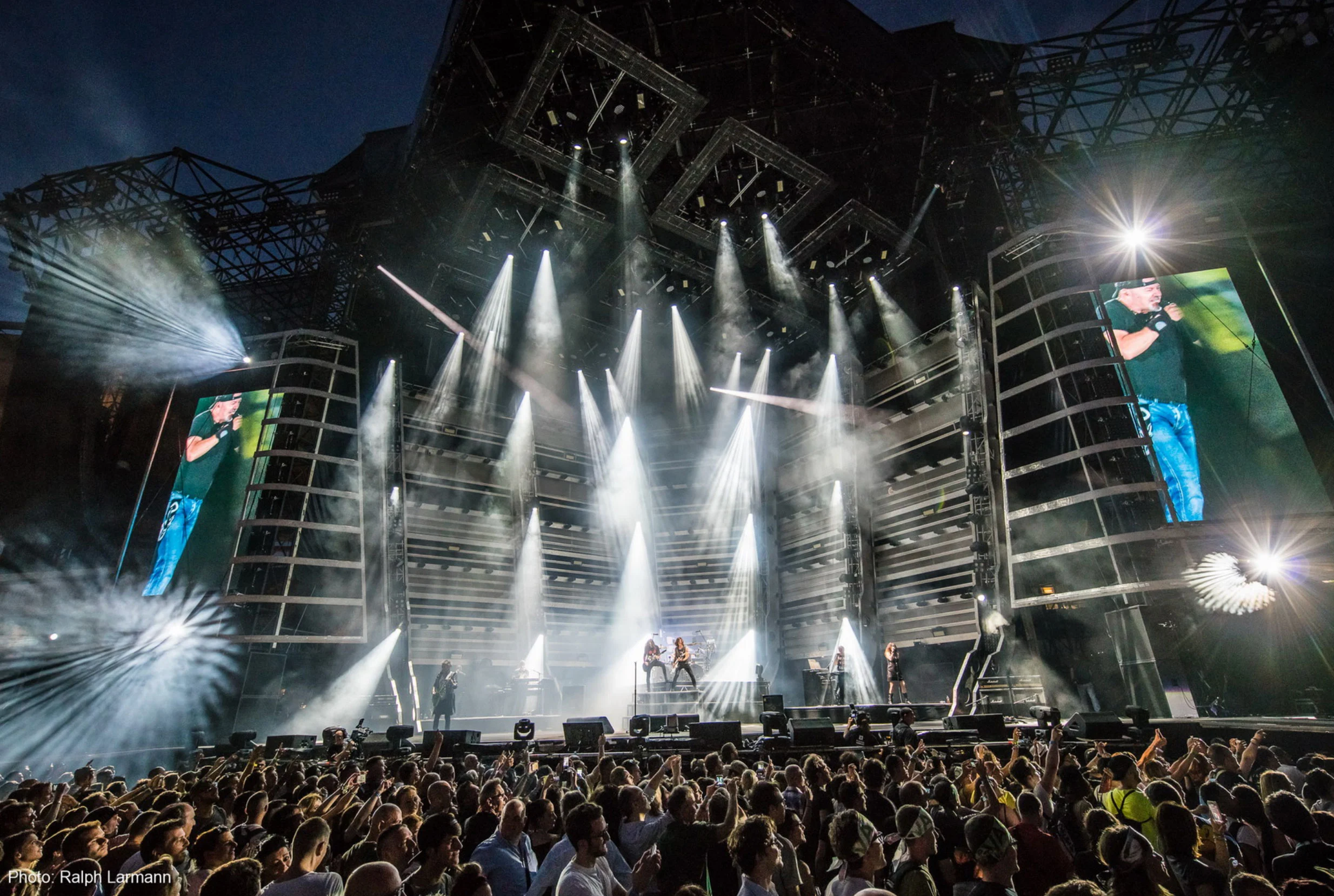 Palco negli stadi di Vasco Live Kom con fasci di luce bianca, struttura angolare di americane e schermi video laterali – Vasco Rossi Live Kom 2016, Italia – scenografia per concerti