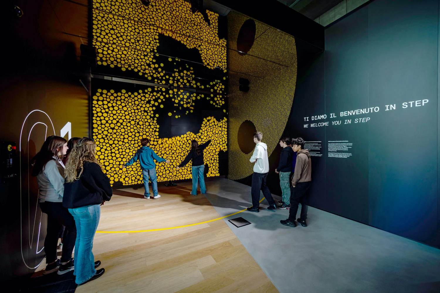 Visitatori davanti a una parete interattiva di punti gialli che reagiscono alle loro sagome, accanto a un pannello di benvenuto – STEP FuturAbility District by Fastweb, Milano, Italia, 2022 – allestimento espositivo
