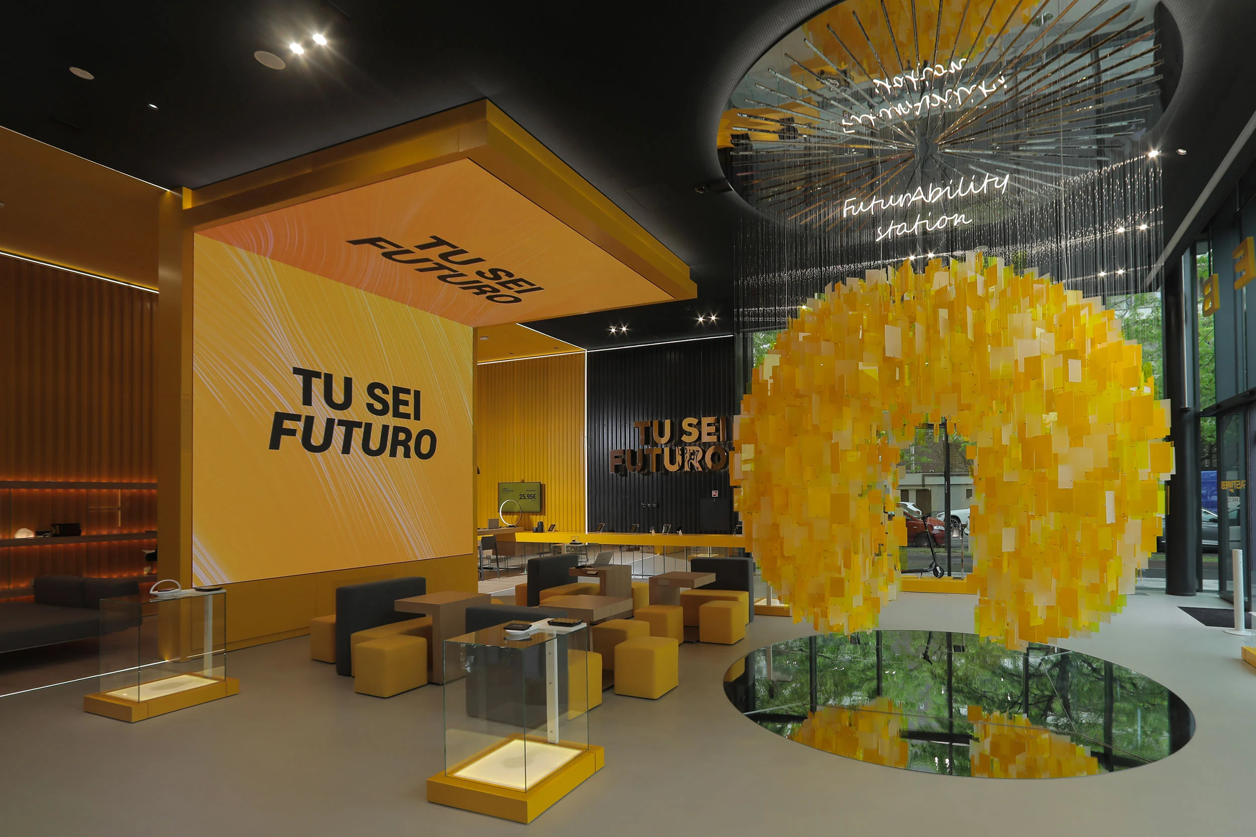 Atrio d'ingresso con schermi gialli "Tu sei futuro", sedute lounge e una sfera sospesa di tessere gialle sopra uno specchio – STEP FuturAbility District by Fastweb, Milano, Italia, 2022 – allestimento espositivo