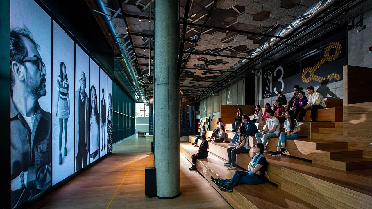 Giovani visitatori su gradonate in legno davanti a una parete di grandi schermi con ritratti in bianco e nero – STEP FuturAbility District by Fastweb, Milano, Italia, 2022 – allestimento espositivo