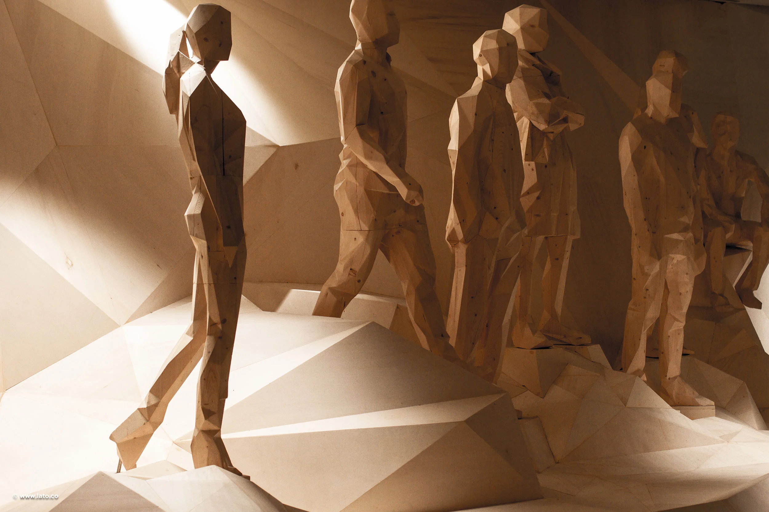 Fila di sculture umane sfaccettate in legno che avanzano su un terreno spigoloso in compensato, a luce radente – Padiglione Italia all'Expo 2015, Milano, Italia – allestimento espositivo