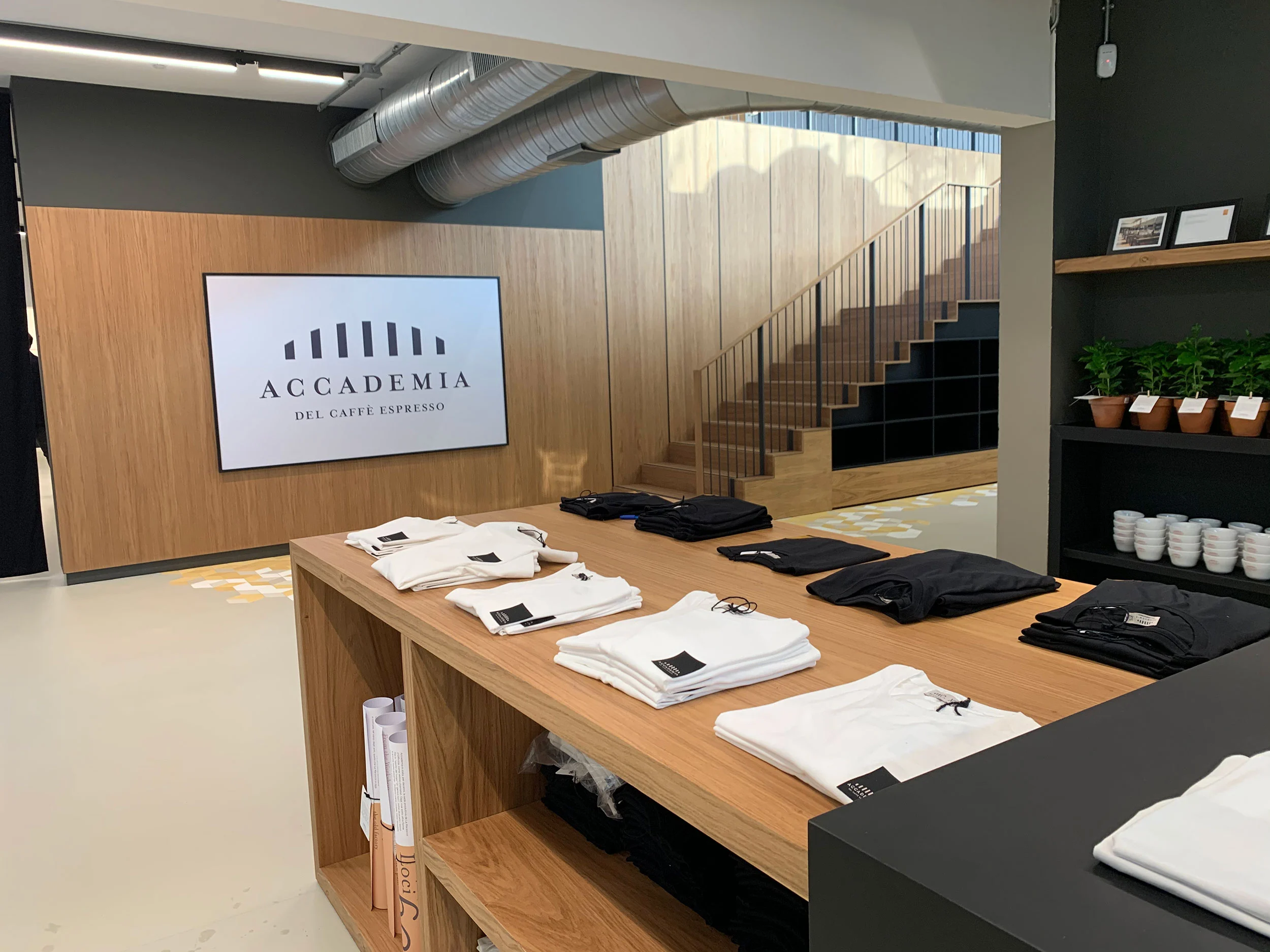 Negozio con magliette piegate su un tavolo di rovere, uno schermo con il logo dell'Accademia e una scala in legno – La Marzocco Accademia del Caffè Espresso, Firenze, Italia, 2018 – interior design