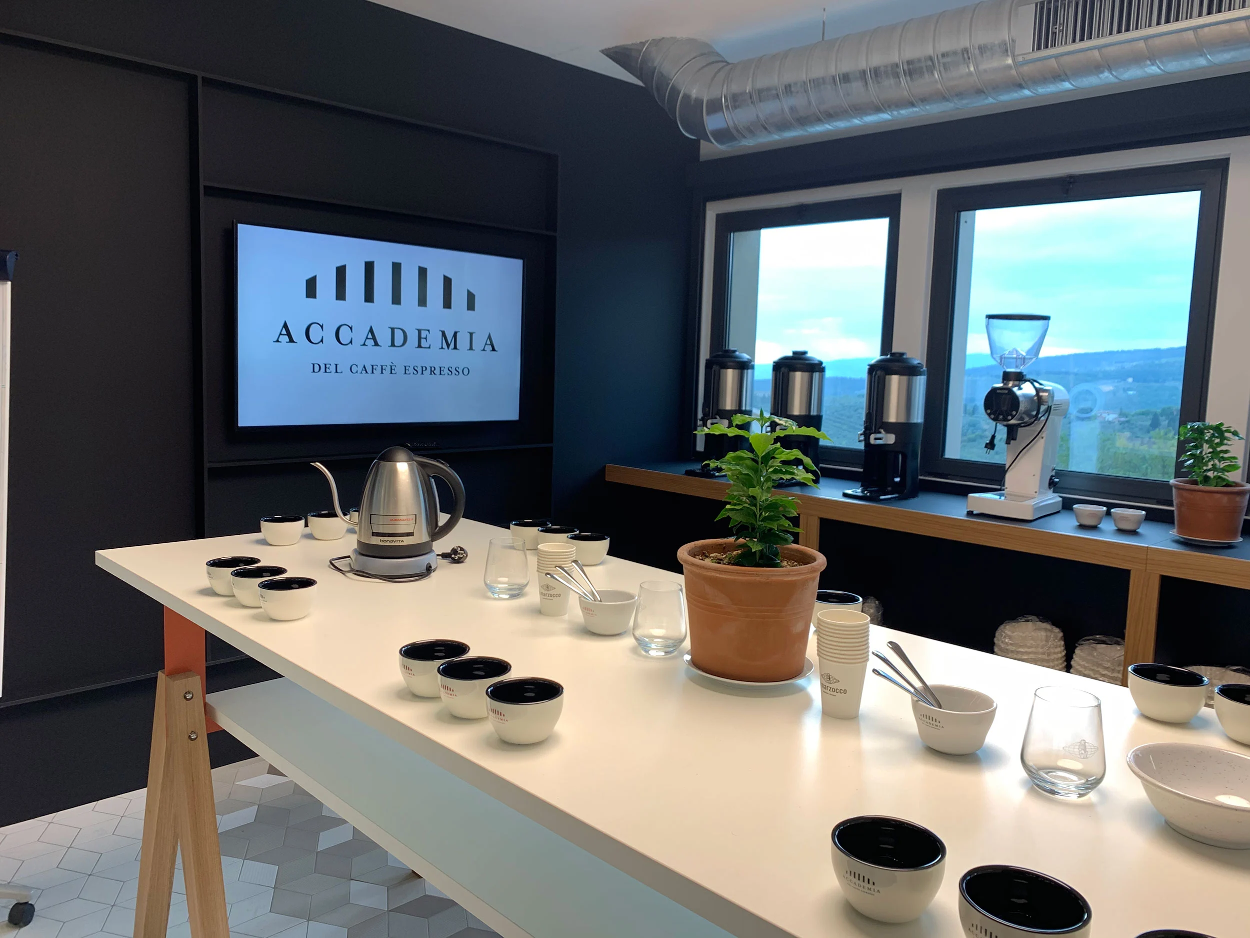 Aula di formazione con un lungo tavolo bianco preparato per il cupping, macinacaffè alle finestre sulle colline – La Marzocco Accademia del Caffè Espresso, Firenze, Italia, 2018 – interior design