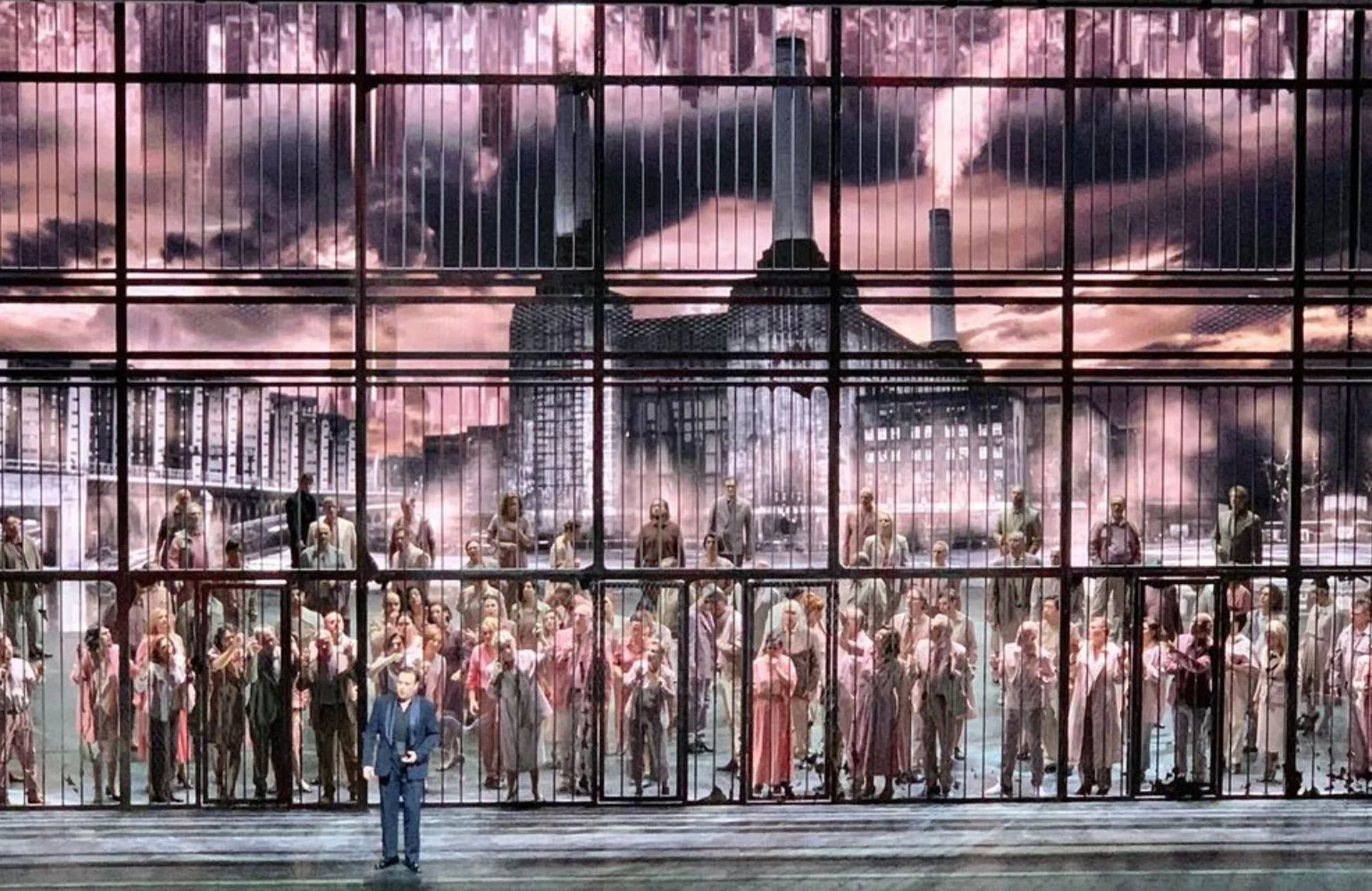 Coro dietro un'alta grata di sbarre, una centrale elettrica fumante proiettata sotto cieli rosa – Macbeth al Teatro alla Scala, Milano, Italia, 2021 – scenografia d'opera