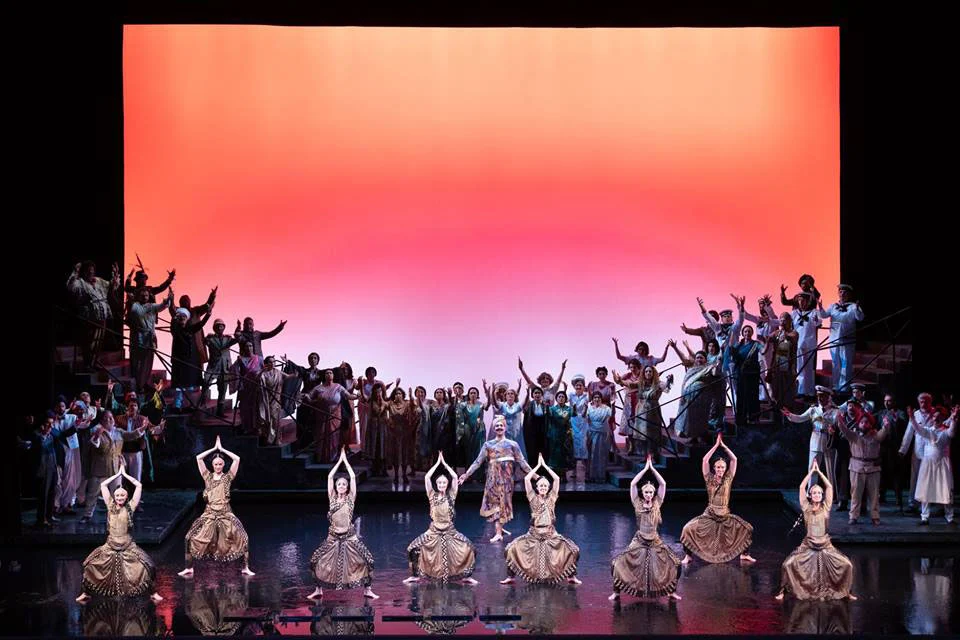 Danzatori in costumi dorati inginocchiati in fila davanti al coro, sotto un vasto fondale rosa-arancio – Lakmé alla Royal Opera House Muscat, Oman, 2019 – scenografia d'opera