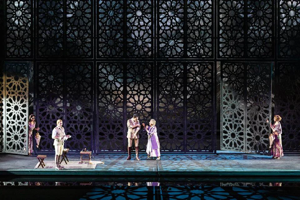 Cantanti davanti ad alti schermi neri con motivi geometrici a traforo, riflessi nel pavimento bagnato del palco – Lakmé alla Royal Opera House Muscat, Oman, 2019 – scenografia d'opera