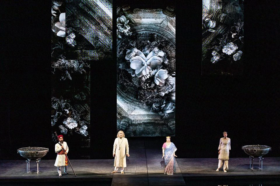 Quattro cantanti su un palco buio tra due vasche in pietra, con proiezioni floreali in bianco e nero sopra – Lakmé alla Royal Opera House Muscat, Oman, 2019 – scenografia d'opera