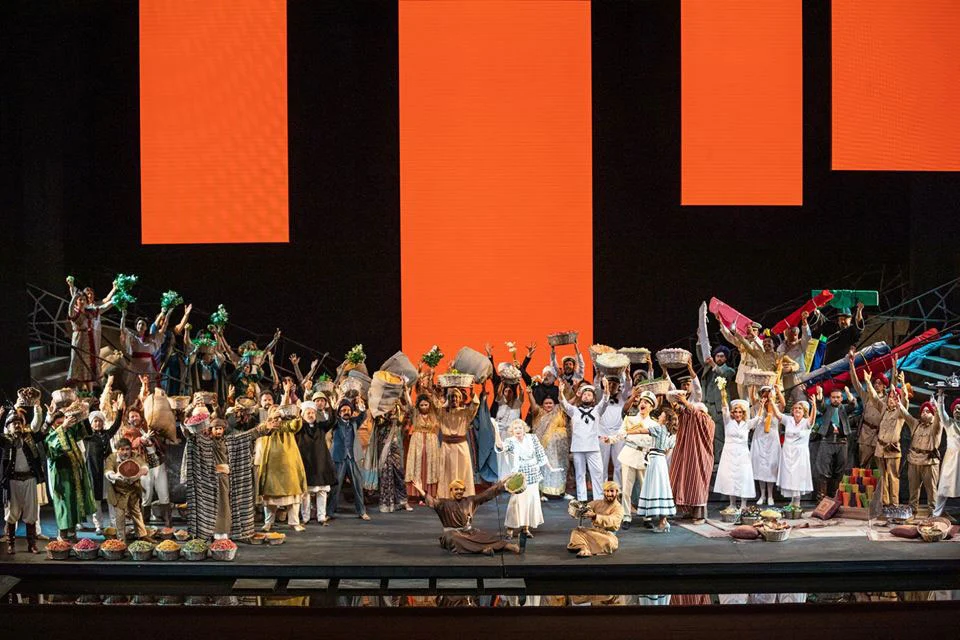 Affollata scena di mercato con il coro che regge cesti e merci sotto grandi pannelli arancioni – Lakmé alla Royal Opera House Muscat, Oman, 2019 – scenografia d'opera