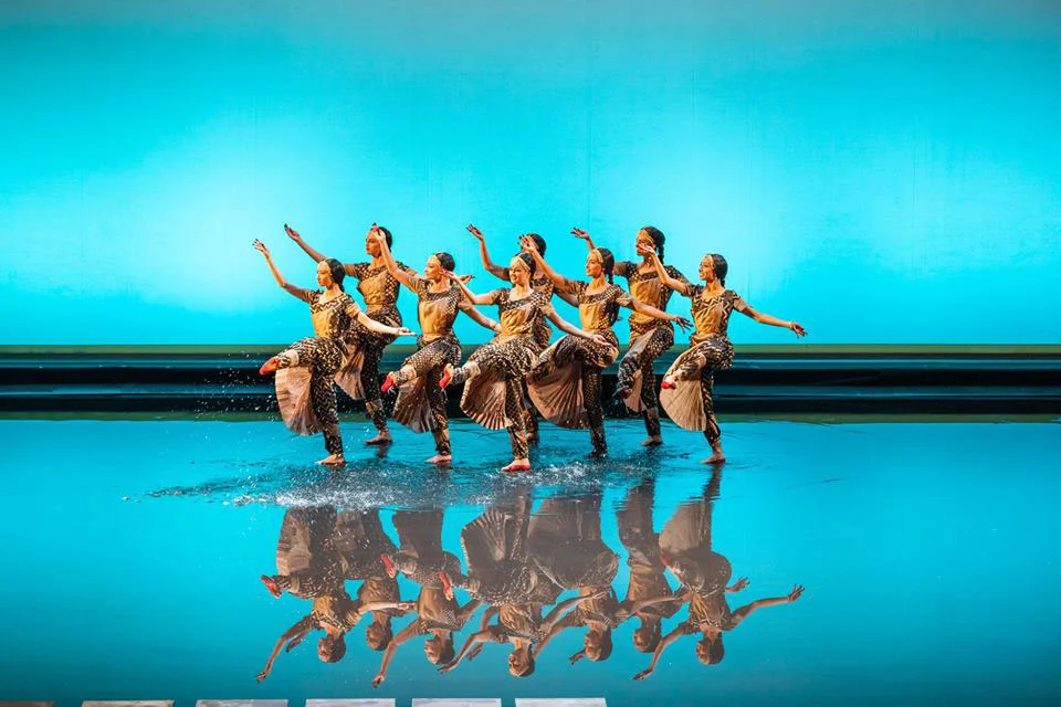 Danzatori in costumi decorati che sguazzano su un palco coperto d'acqua, riflessi contro un fondale turchese – Lakmé alla Royal Opera House Muscat, Oman, 2019 – scenografia d'opera
