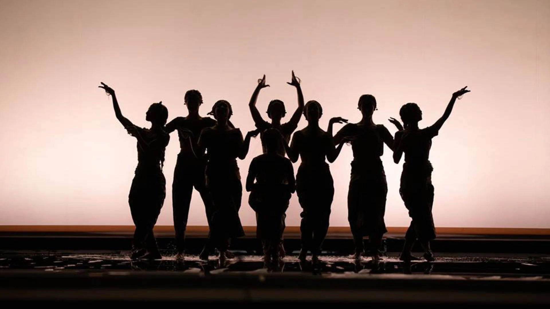 Fila di danzatori in controluce a braccia alzate davanti a un fondale rosa tenuemente luminoso – Lakmé alla Royal Opera House Muscat, Oman, 2019 – scenografia d'opera
