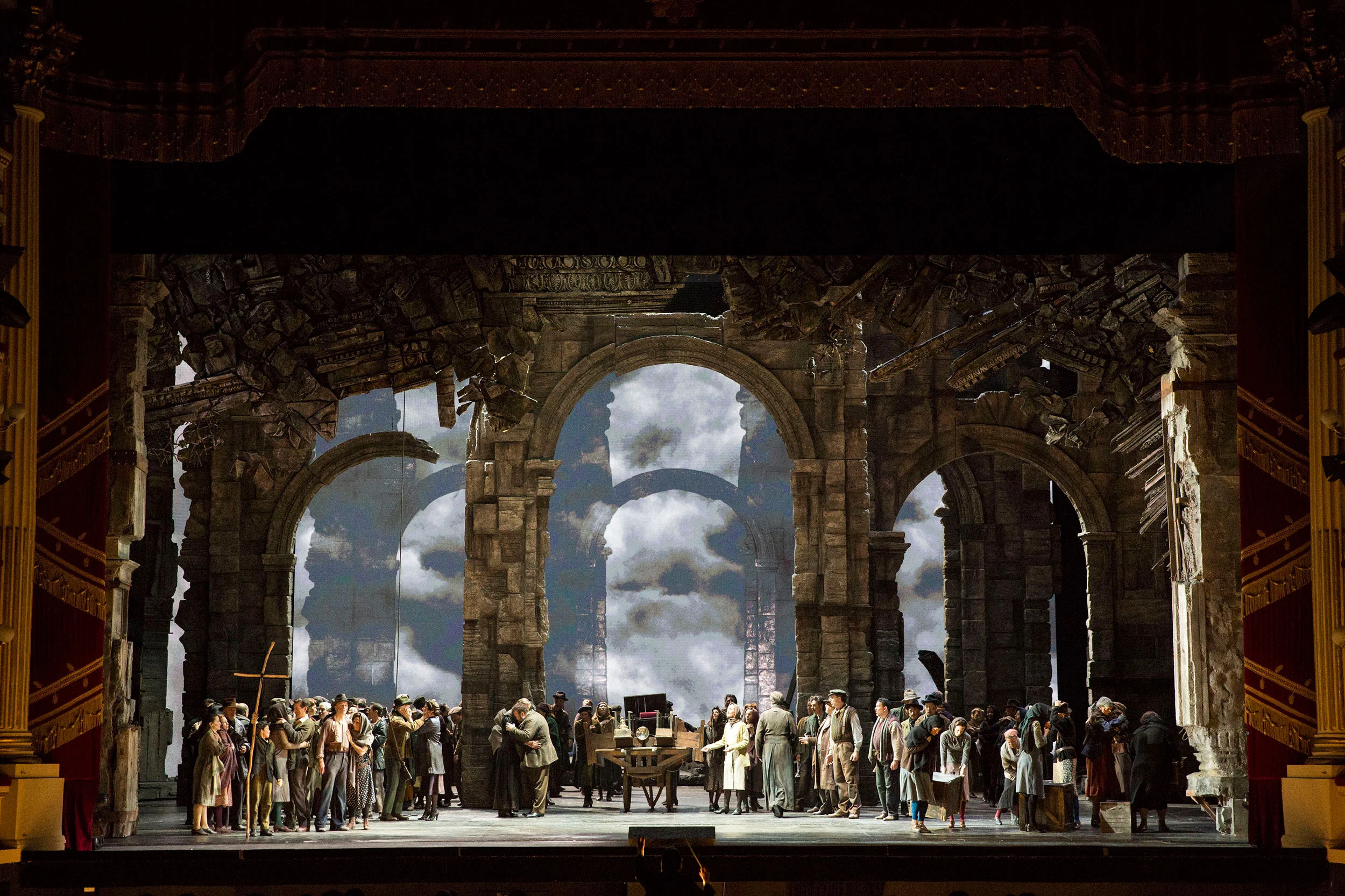 Folla di paesani in un'ambientazione romana in rovina, con archi di pietra aperti su un cielo nuvoloso – Attila al Teatro alla Scala, Milano, Italia, 2018 – scenografia d'opera