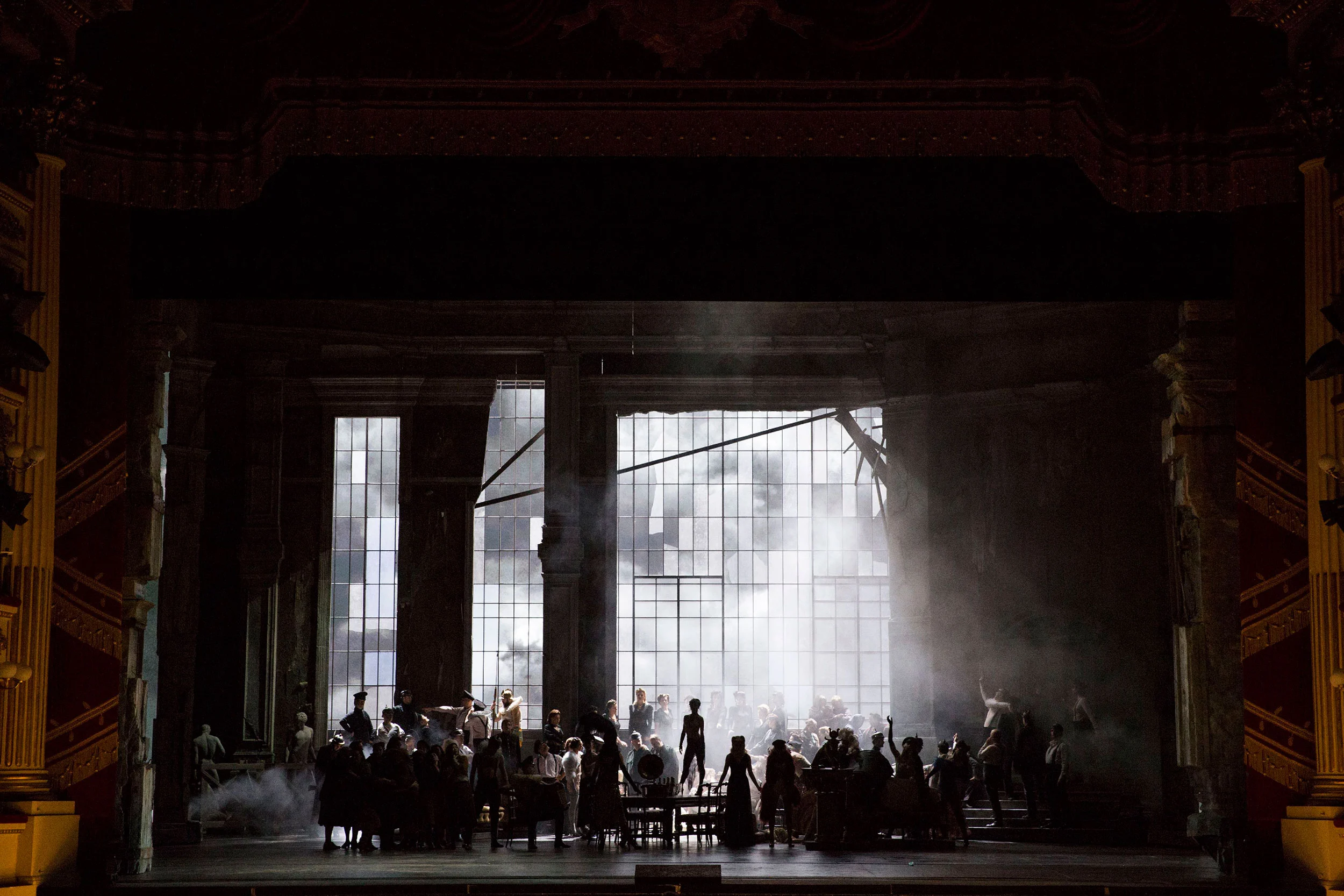 Folla in controluce davanti a una vetrata infranta luminosa, fumo che attraversa la sala buia – Attila al Teatro alla Scala, Milano, Italia, 2018 – scenografia d'opera