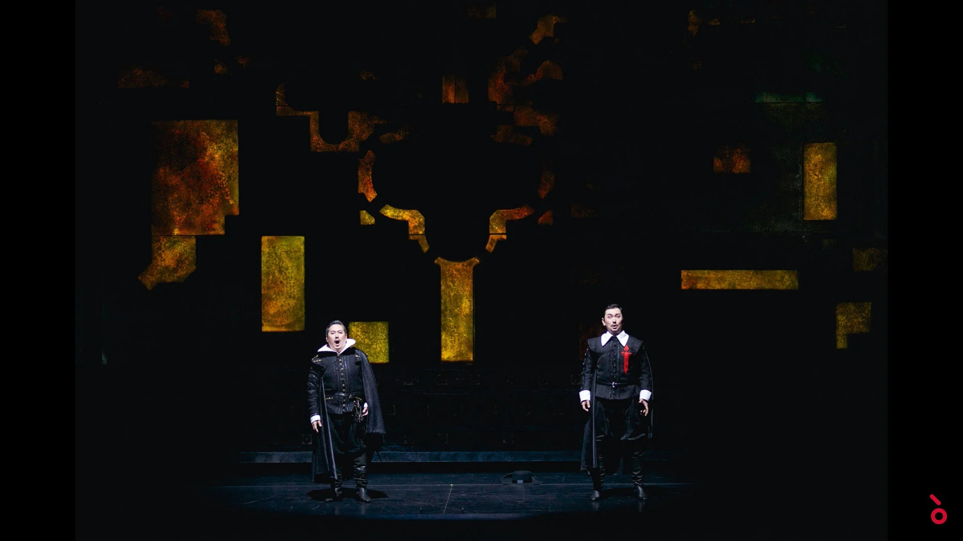 Due cantanti in costume spagnolo nero su un palco buio con frammenti dorati di una croce dietro – Don Carlo all'Astana Opera, Kazakistan, 2023 – scenografia d'opera