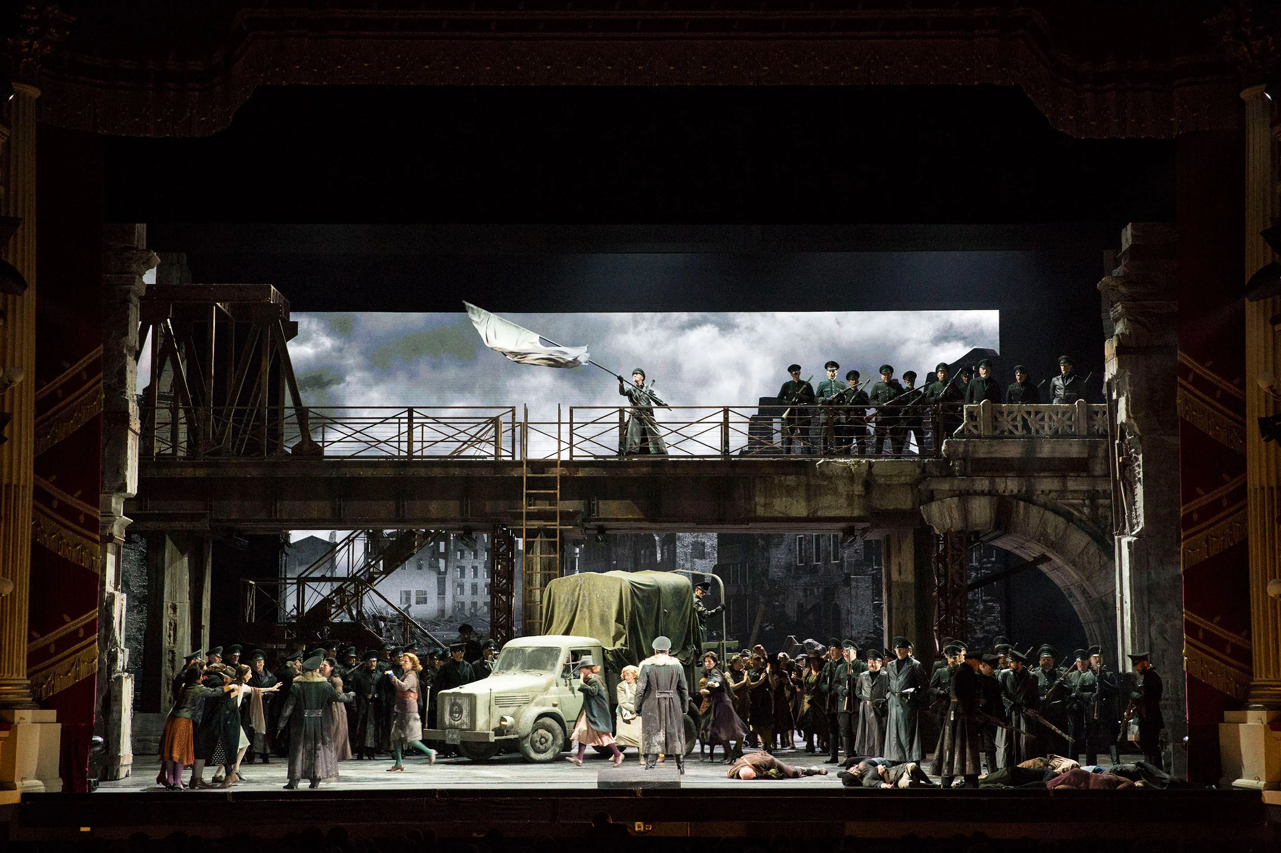 Ponte monumentale con soldati e una bandiera al vento sopra una folla e un camion militare – Attila al Teatro alla Scala, Milano, Italia, 2018 – scenografia d'opera
