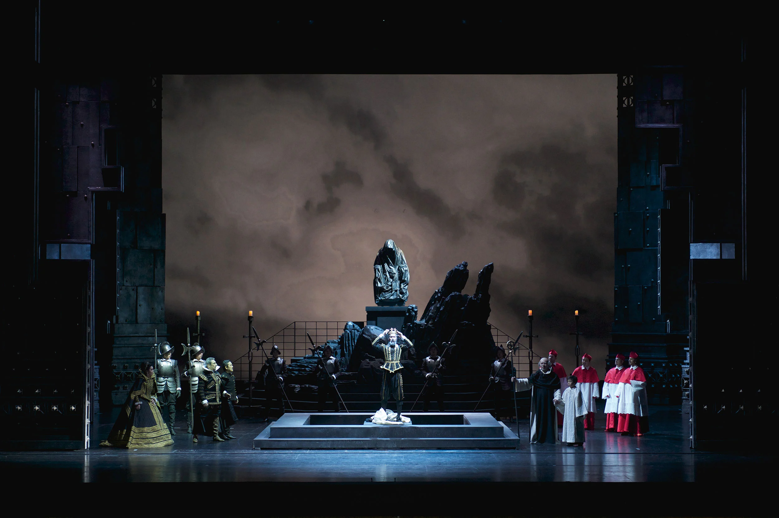 Cantante inginocchiato su una pedana davanti a una statua in pietra, cardinali e soldati ai lati sotto un cielo nuvoloso – Don Carlo all'Astana Opera, Kazakistan, 2023 – scenografia d'opera