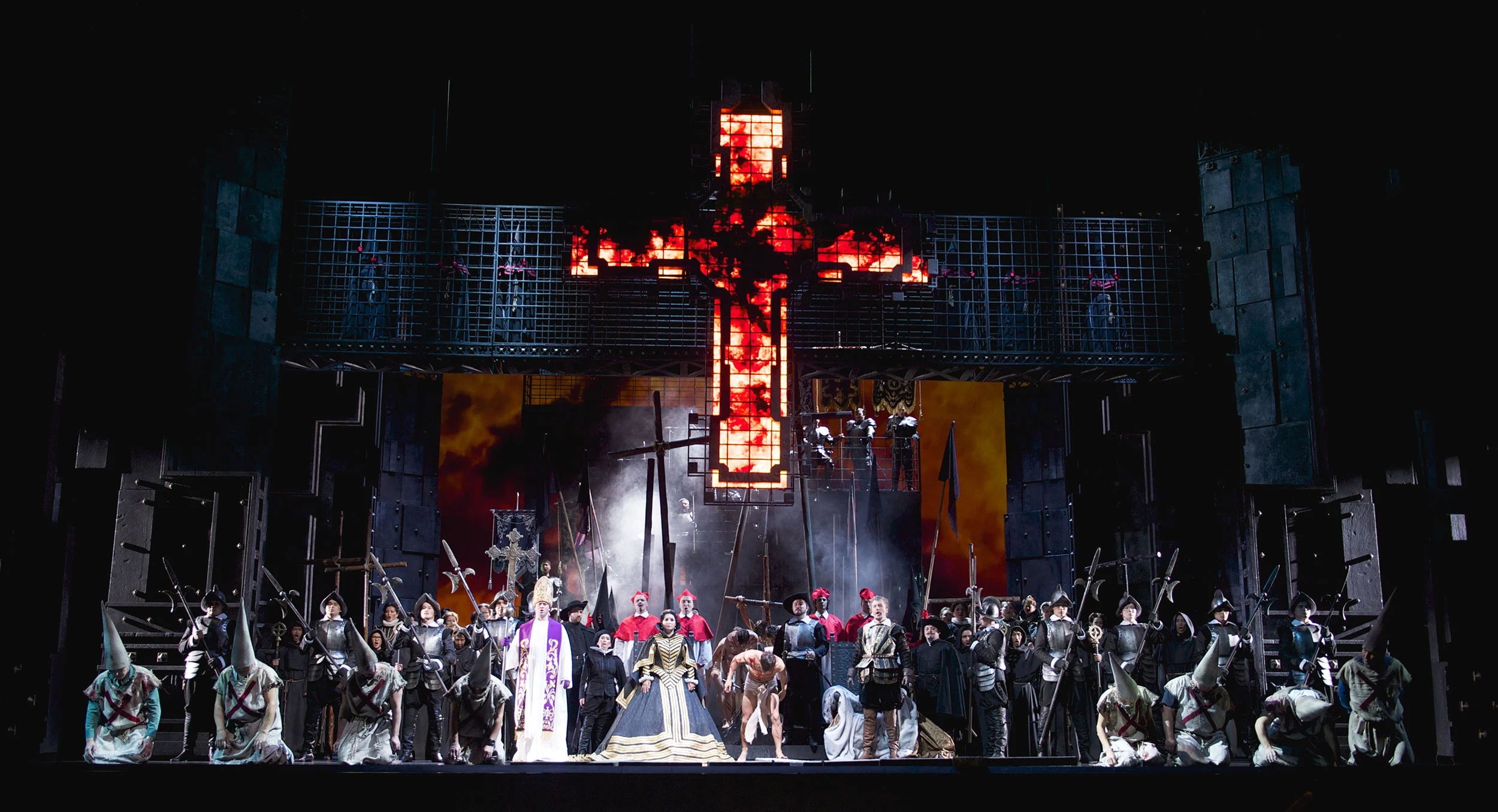 Finale con tutto il cast davanti a una croce in fiamme, con penitenti incappucciati davanti a una griglia di gabbie di ferro – Don Carlo all'Astana Opera, Kazakistan, 2023 – scenografia d'opera