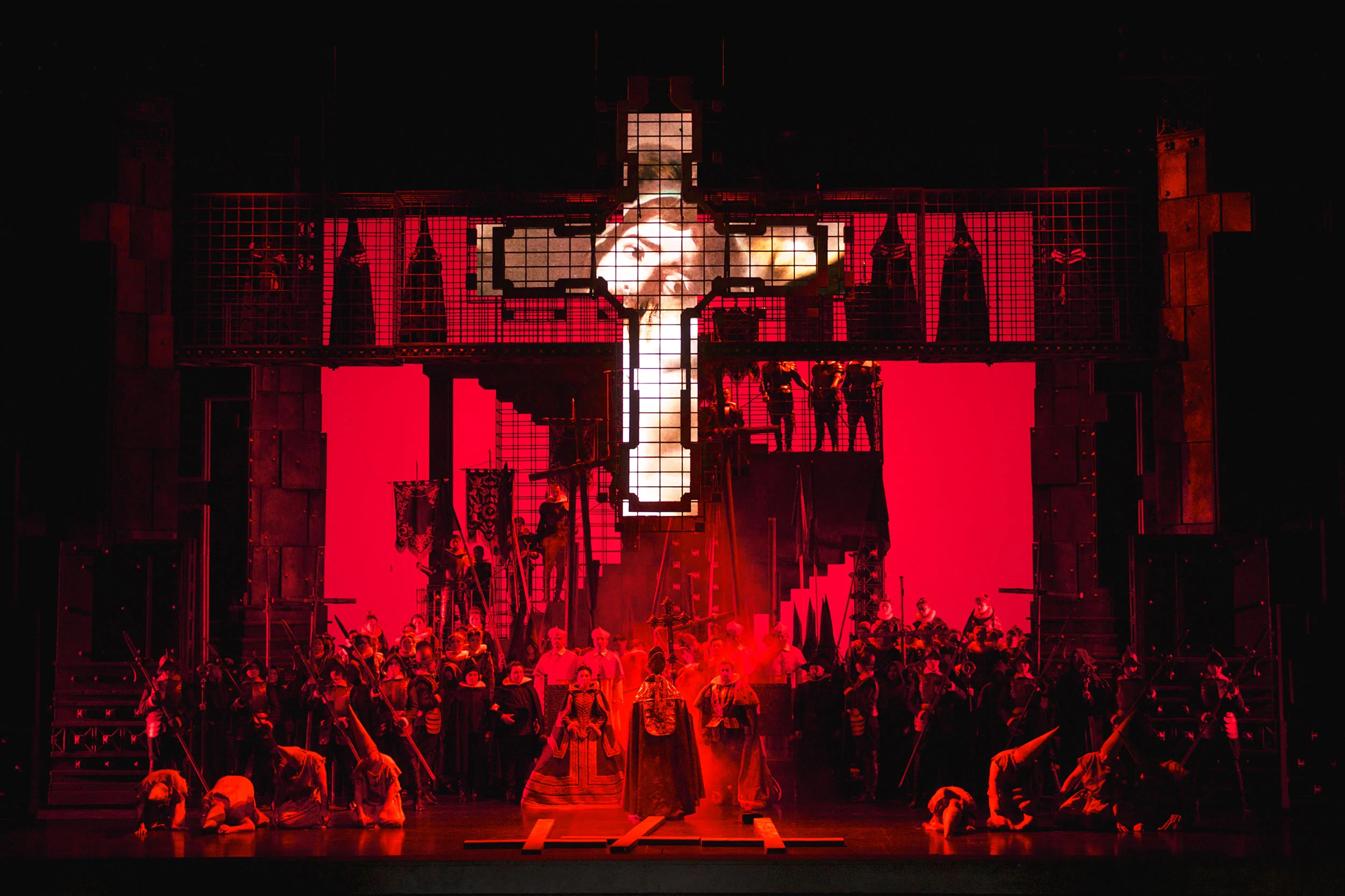 Palco inondato di luce rossa sotto una gigantesca croce di luce, il coro schierato davanti alla struttura delle gabbie – Don Carlo all'Astana Opera, Kazakistan, 2023 – scenografia d'opera