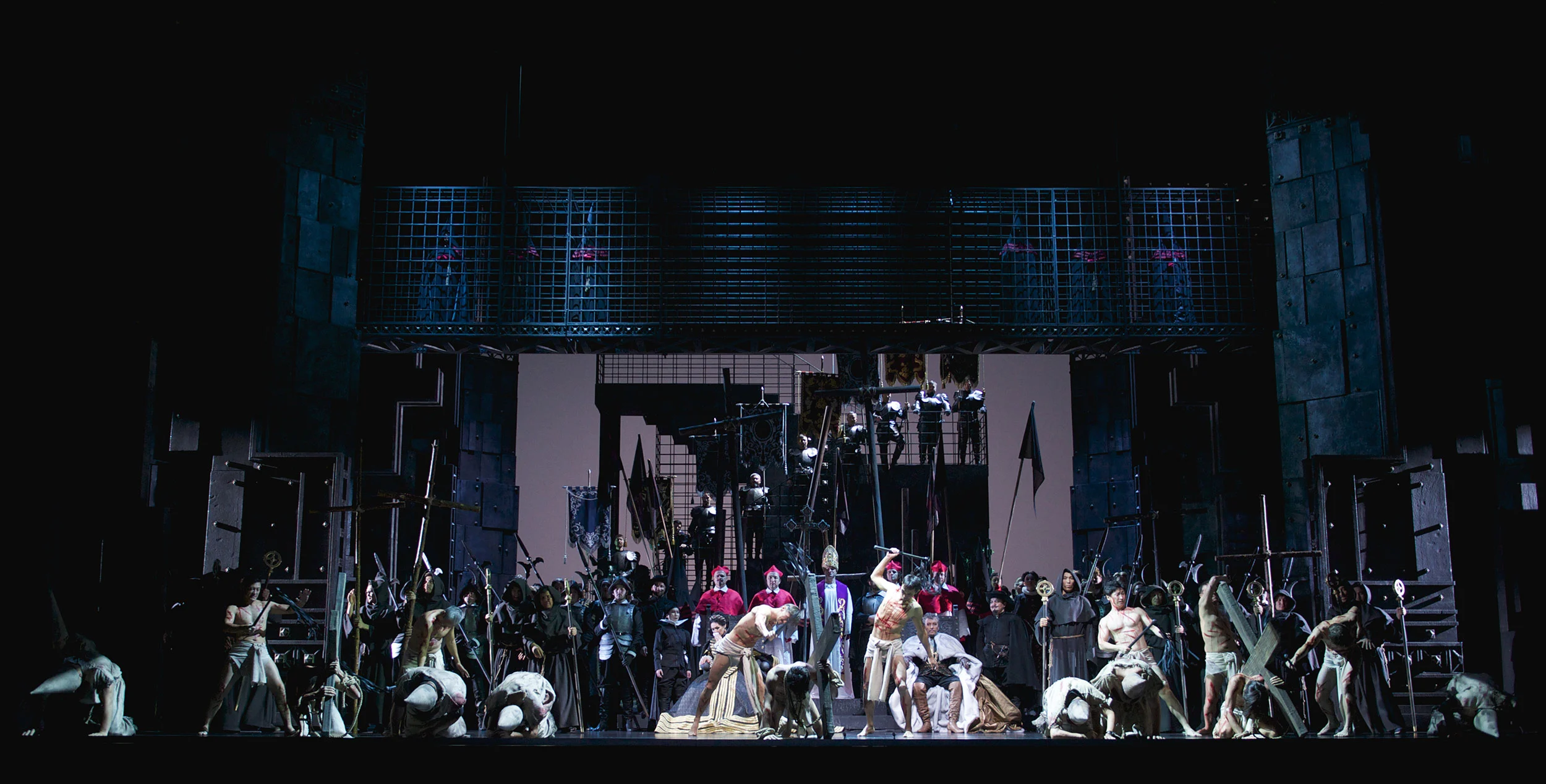 Vista d'insieme di una scena di massa con bandiere e soldati sotto le gabbie di ferro, figure inginocchiate davanti – Don Carlo all'Astana Opera, Kazakistan, 2023 – scenografia d'opera