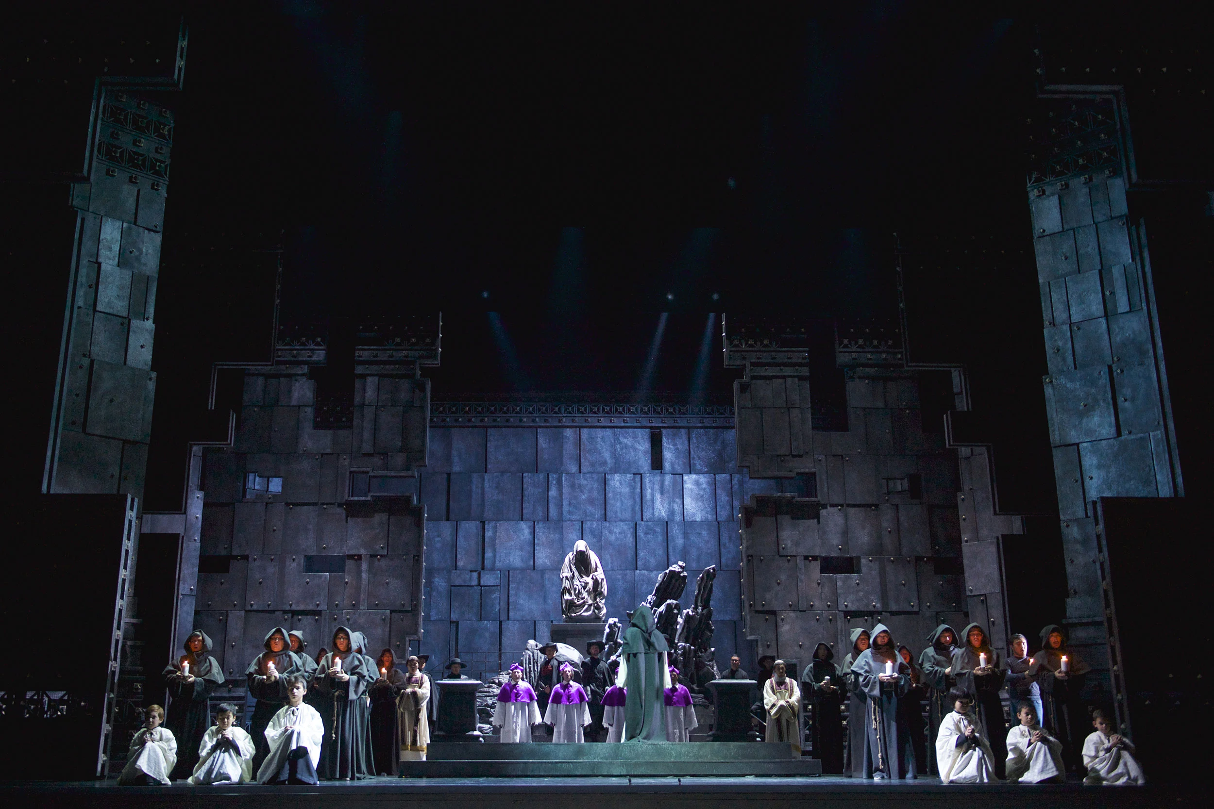 Monaci con candele e una statua al centro davanti a una scenografia di un monumentale muro in pietra grigia – Don Carlo all'Astana Opera, Kazakistan, 2023 – scenografia d'opera