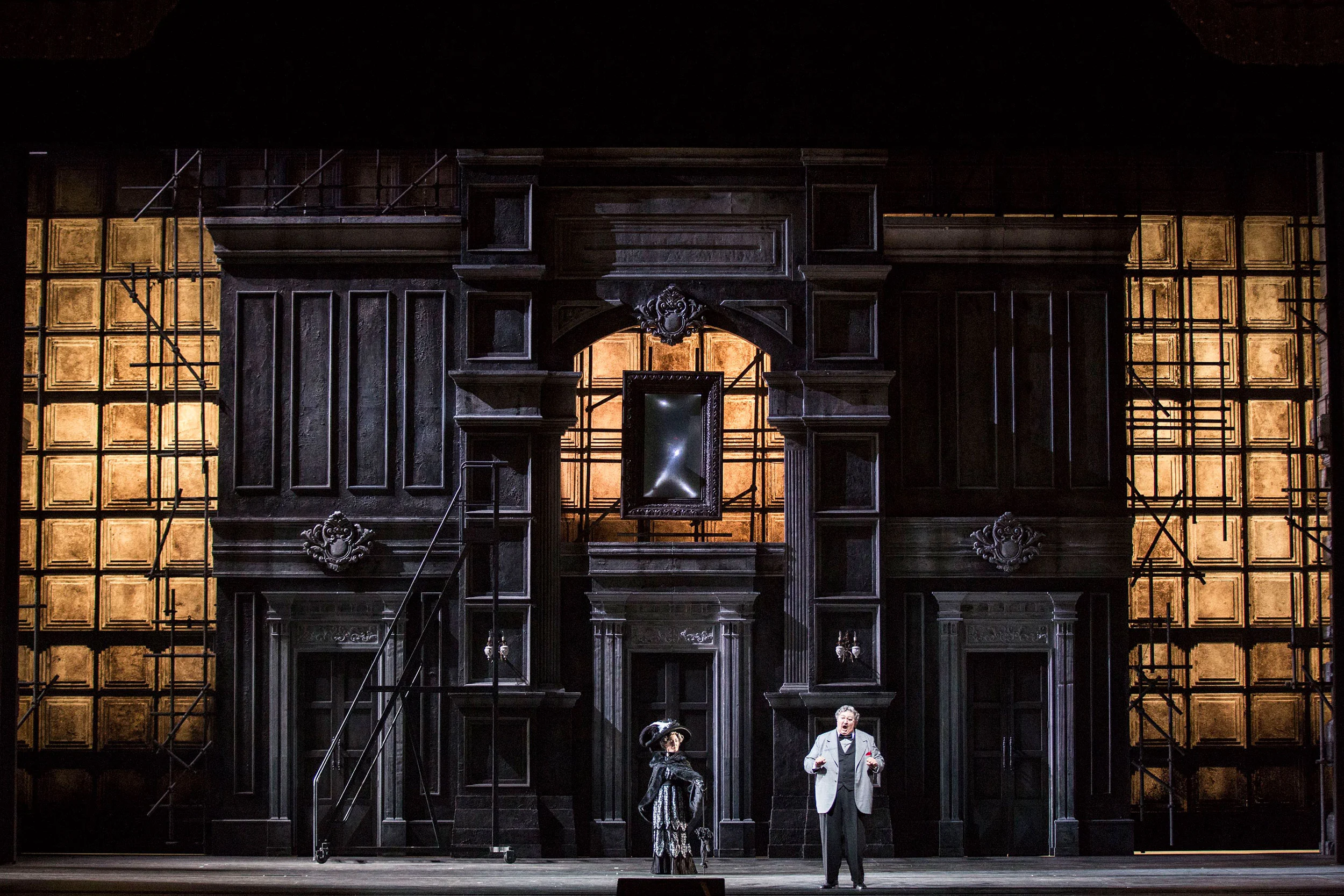 Due cantanti davanti a una facciata grigia scolpita incorniciata da ponteggi, con finestre dalla luce calda – Don Pasquale al Teatro alla Scala, Milano, Italia, 2018 – scenografia d'opera
