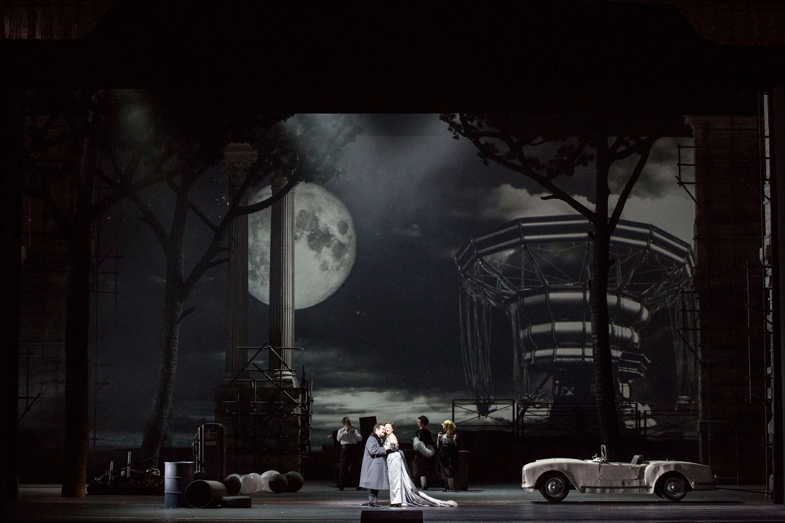 Scena notturna con luna piena, alberi spogli, la struttura di una giostra e una decappottabile bianca d'epoca – Don Pasquale al Teatro alla Scala, Milano, Italia, 2018 – scenografia d'opera