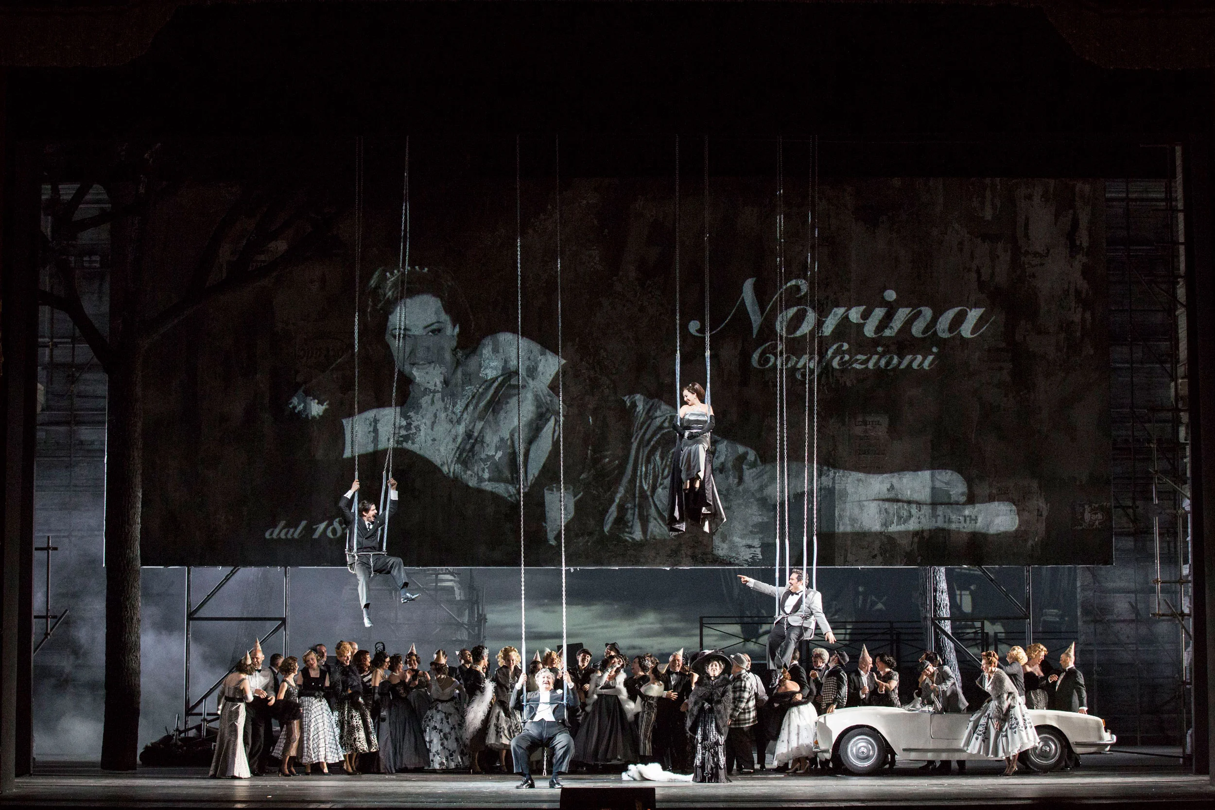 Coro sul palco con altalene appese davanti alla proiezione di una donna e della scritta Norina Confezioni – Don Pasquale al Teatro alla Scala, Milano, Italia, 2018 – scenografia d'opera