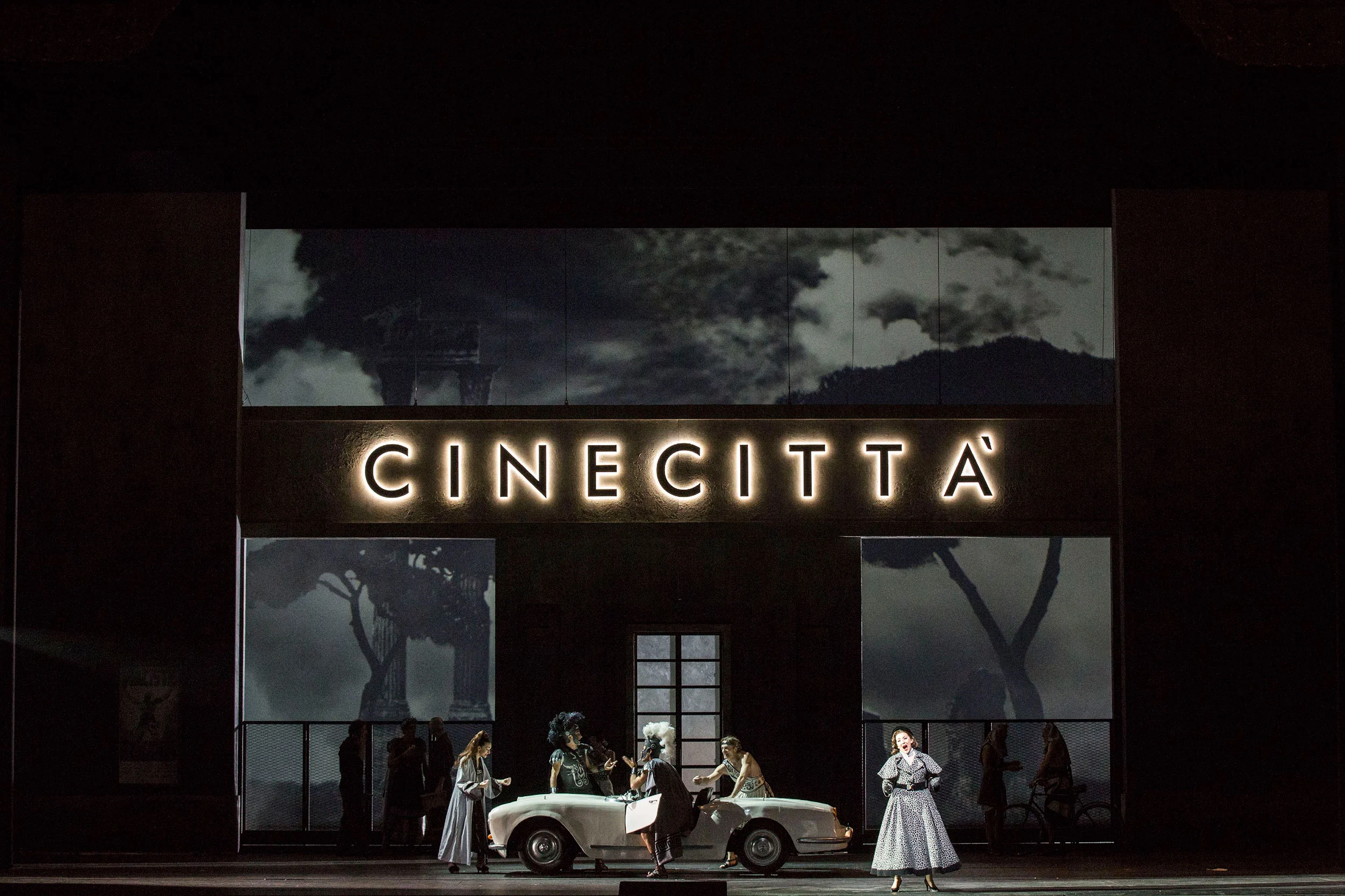 Insegna luminosa di Cinecittà sopra la facciata di un edificio, cantanti intorno a una decappottabile bianca – Don Pasquale al Teatro alla Scala, Milano, Italia, 2018 – scenografia d'opera