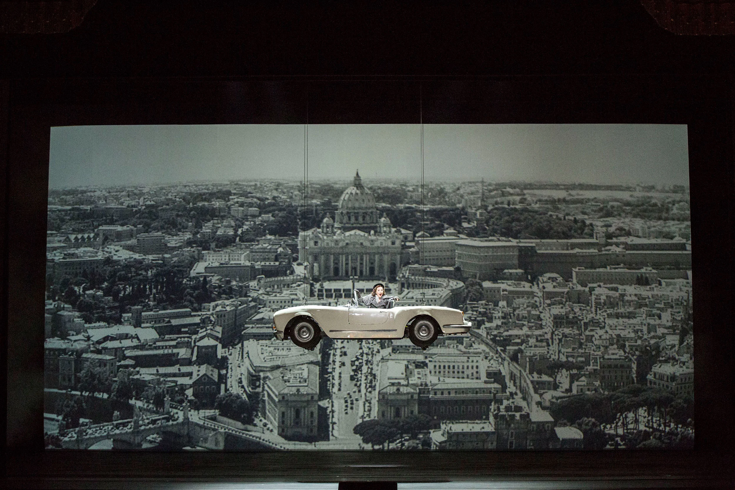 Cantante alla guida di una decappottabile bianca sospesa sopra una vista aerea in bianco e nero di Roma – Don Pasquale al Teatro alla Scala, Milano, Italia, 2018 – scenografia d'opera