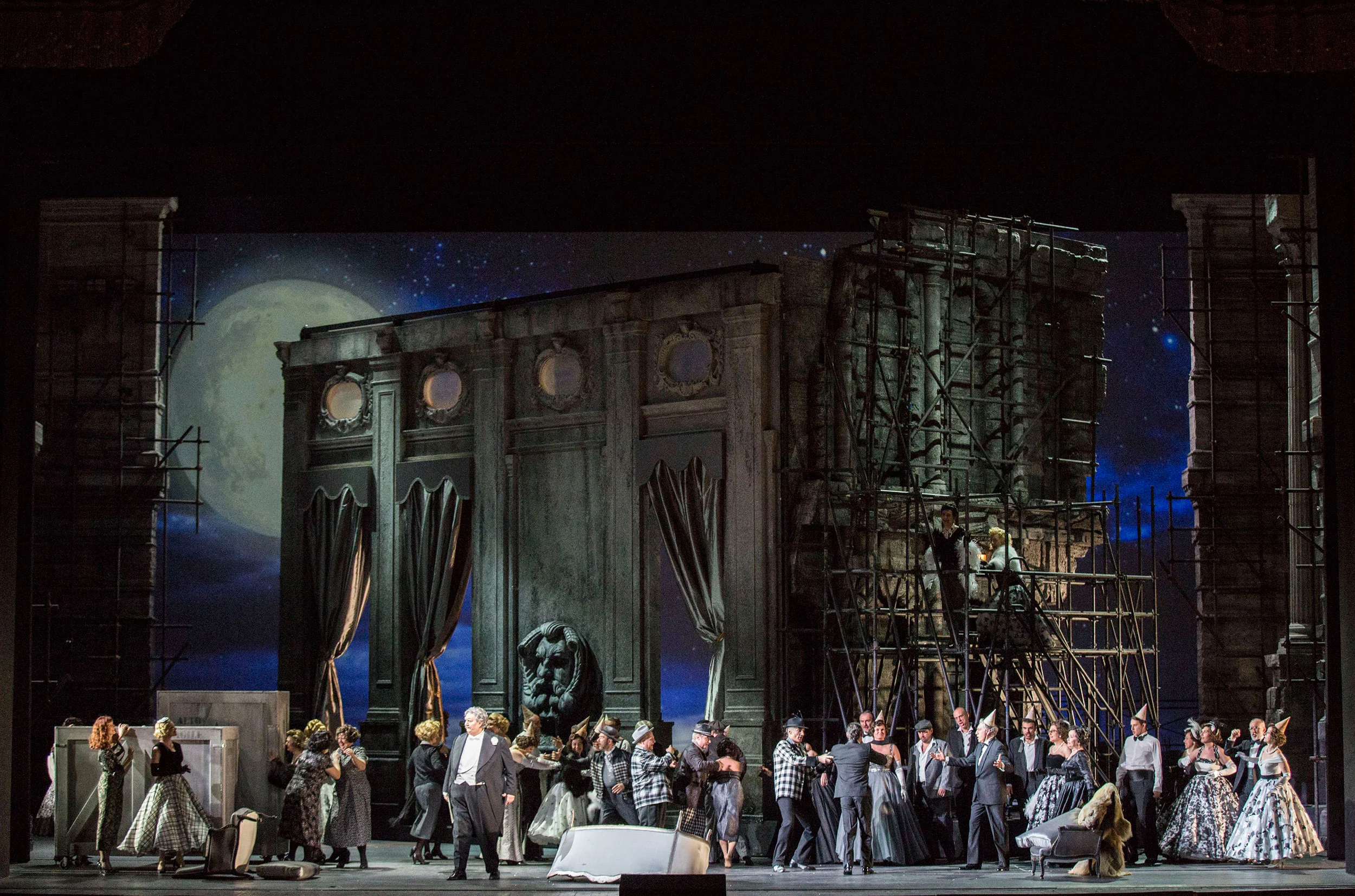 Tutto il cast davanti alla facciata scura di un palazzo e a ponteggi sotto un cielo blu al chiaro di luna – Don Pasquale al Teatro alla Scala, Milano, Italia, 2018 – scenografia d'opera
