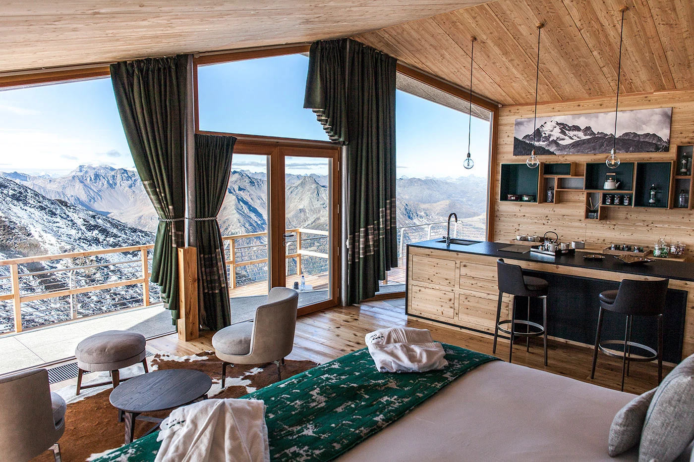Suite dello chalet con letto, poltrone e bancone cucina sotto un soffitto in legno, vetrate a tutta altezza sulle montagne – Levissima 3000 Chalet, Valdisotto, Italia, 2018 – architettura