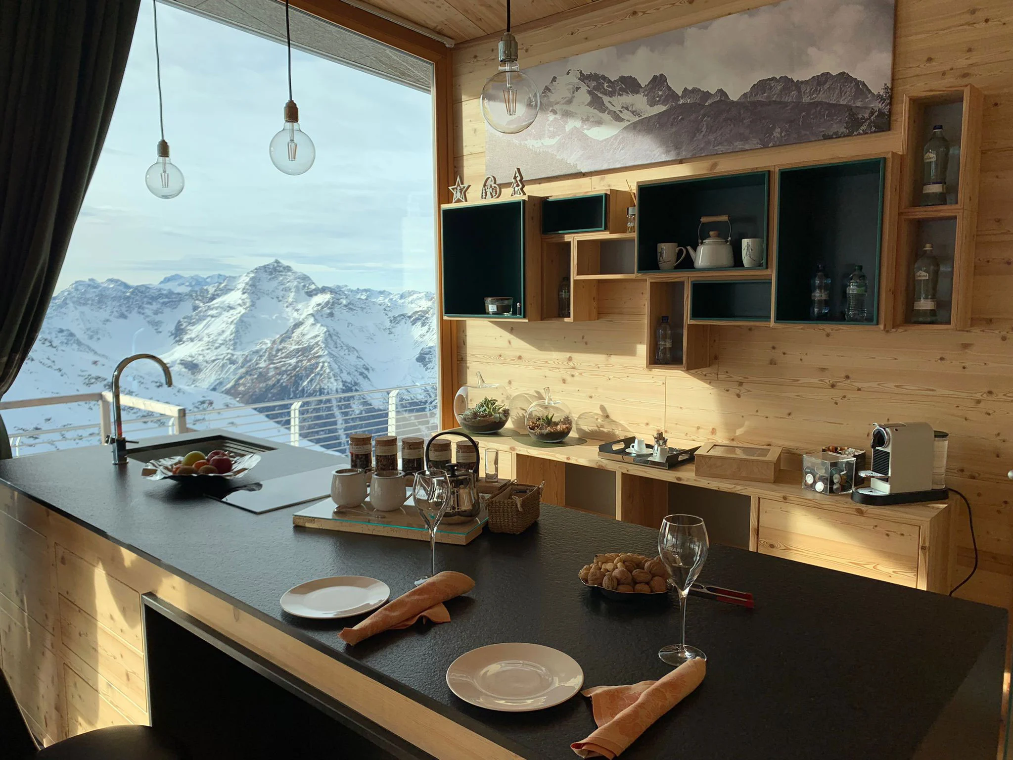 Bancone cucina in legno chiaro e pietra scura apparecchiato per la colazione, con una vetta innevata inquadrata dalla finestra – Levissima 3000 Chalet, Valdisotto, Italia, 2018 – architettura