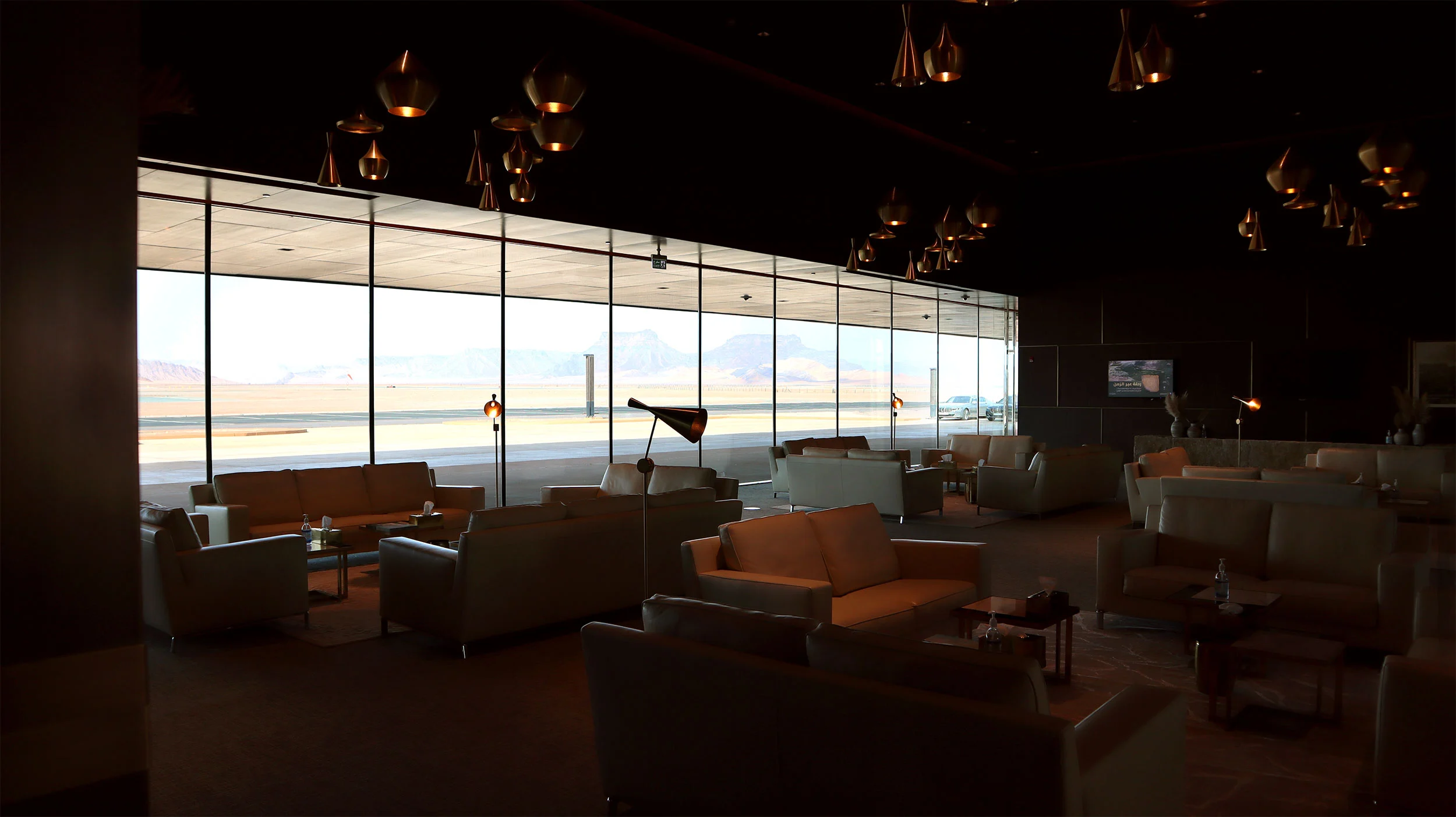 Lounge con divani in pelle e lampade a sospensione in rame, vetrate a tutta altezza sull'aeroporto – Aeroporto Principe Abdul Majeed bin Abdulaziz, AlUla, Arabia Saudita, 2021 – architettura
