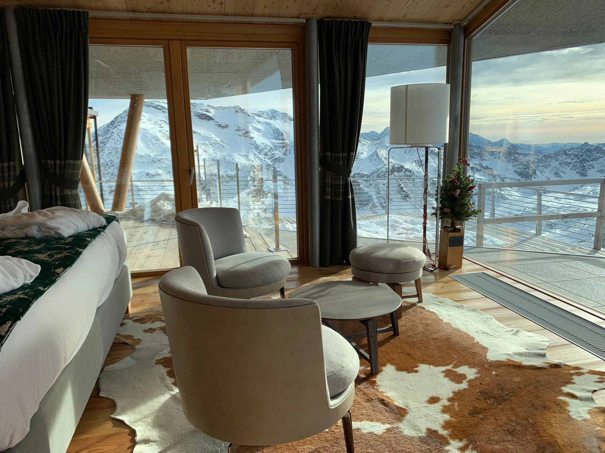 Angolo salotto della camera con poltrone grigie su un tappeto di pelle bovina, vetrate aperte sulla terrazza e sulle Alpi – Levissima 3000 Chalet, Valdisotto, Italia, 2018 – architettura