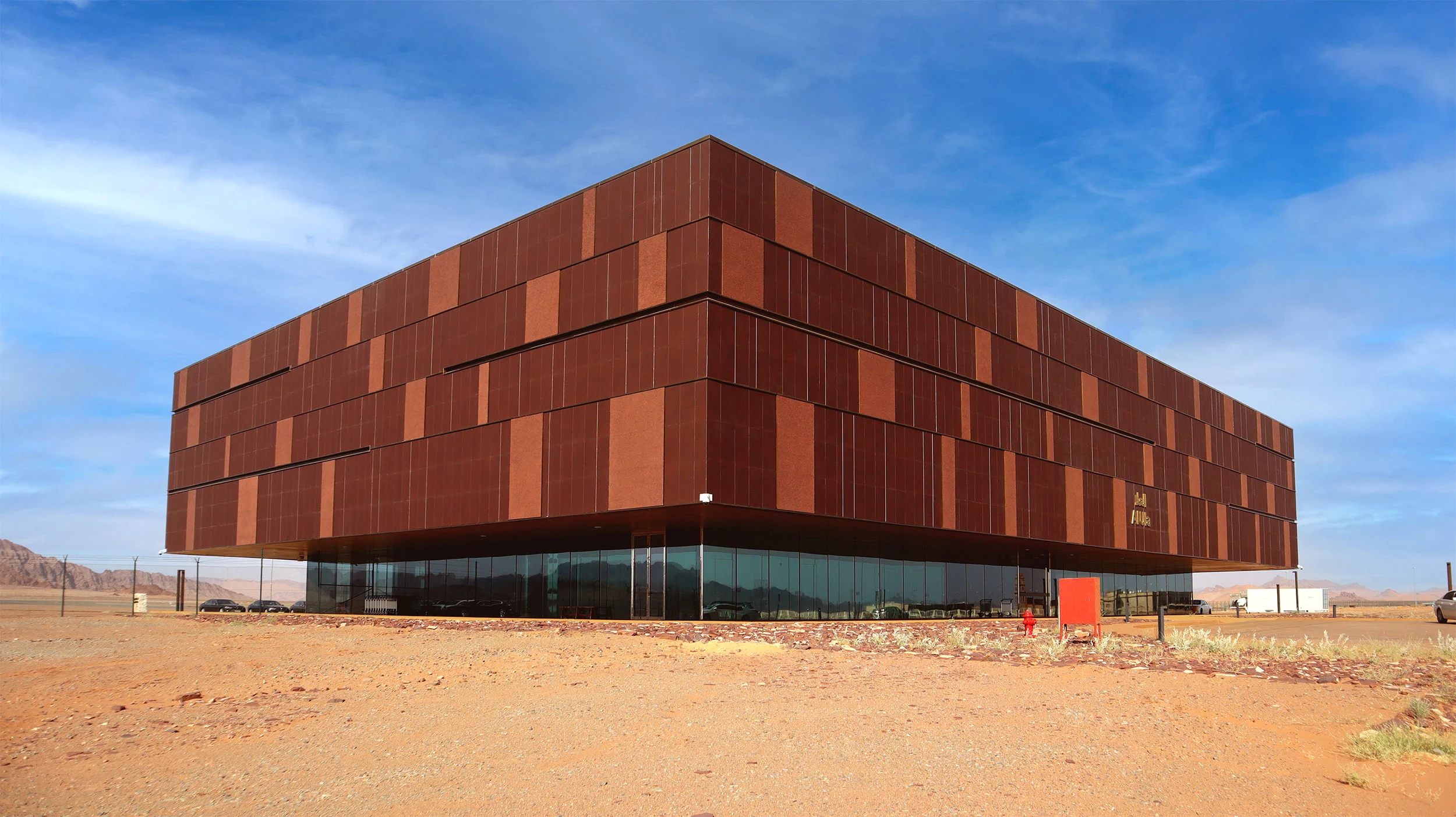 Il volume a sbalzo del terminal rivestito di pannelli rossastri, sospeso sopra una base vetrata nel deserto – Aeroporto Principe Abdul Majeed bin Abdulaziz, AlUla, Arabia Saudita, 2021 – architettura