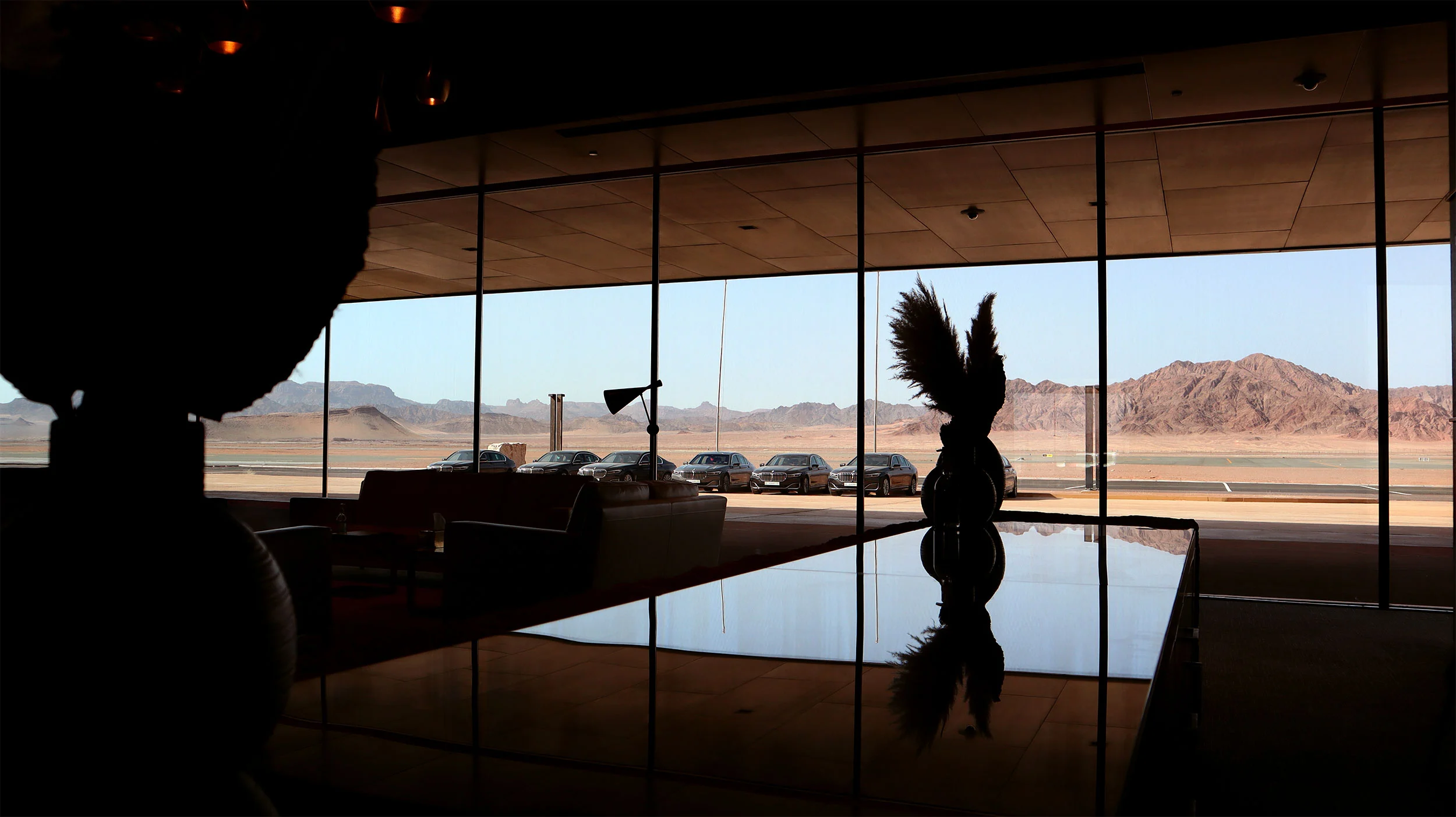 Interno della lounge in controluce, un tavolo lucido che riflette una scultura a palma davanti alla vista sulle montagne – Aeroporto Principe Abdul Majeed bin Abdulaziz, AlUla, Arabia Saudita, 2021 – architettura