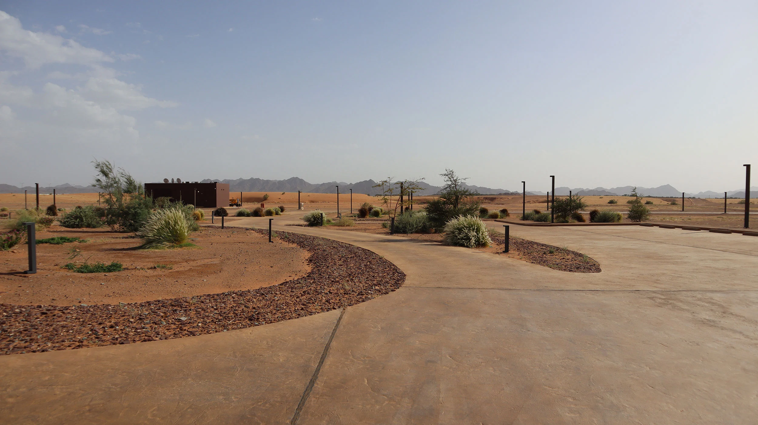 Piazzale d'ingresso paesaggistico con percorsi curvi, aiuole in ghiaia e piante del deserto, montagne all'orizzonte – Aeroporto Principe Abdul Majeed bin Abdulaziz, AlUla, Arabia Saudita, 2021 – architettura