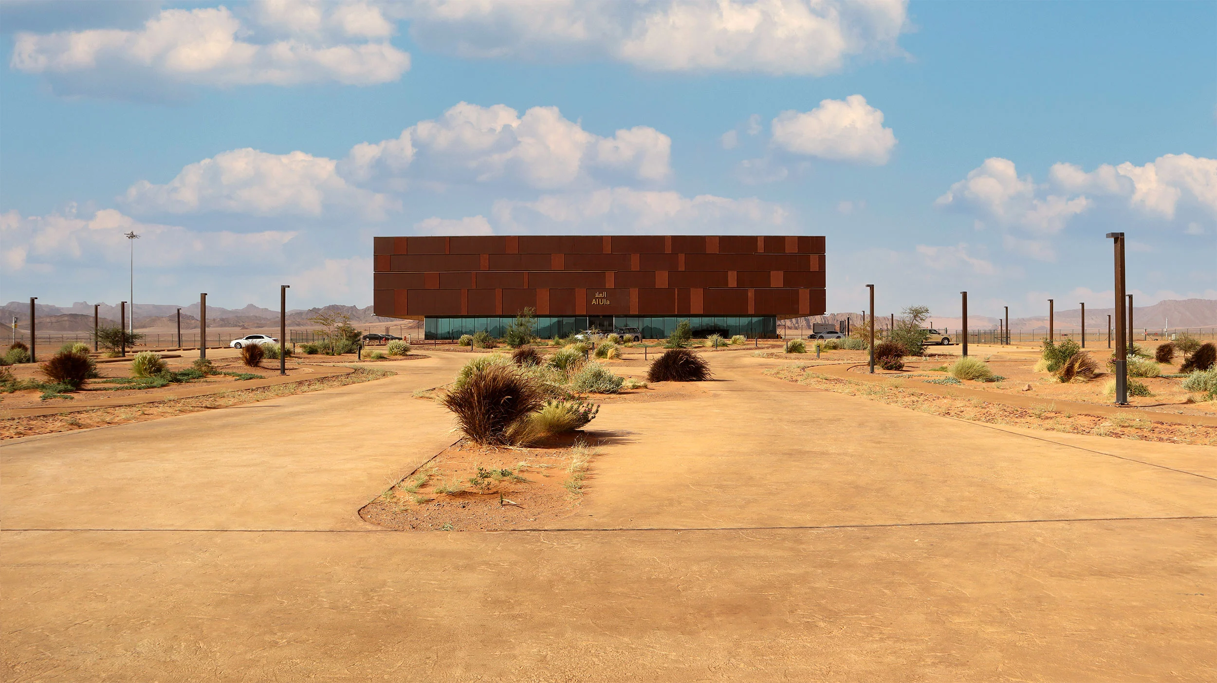 Vista frontale da lontano del terminal oltre una strada d'accesso curva nel deserto bordata di arbusti – Aeroporto Principe Abdul Majeed bin Abdulaziz, AlUla, Arabia Saudita, 2021 – architettura