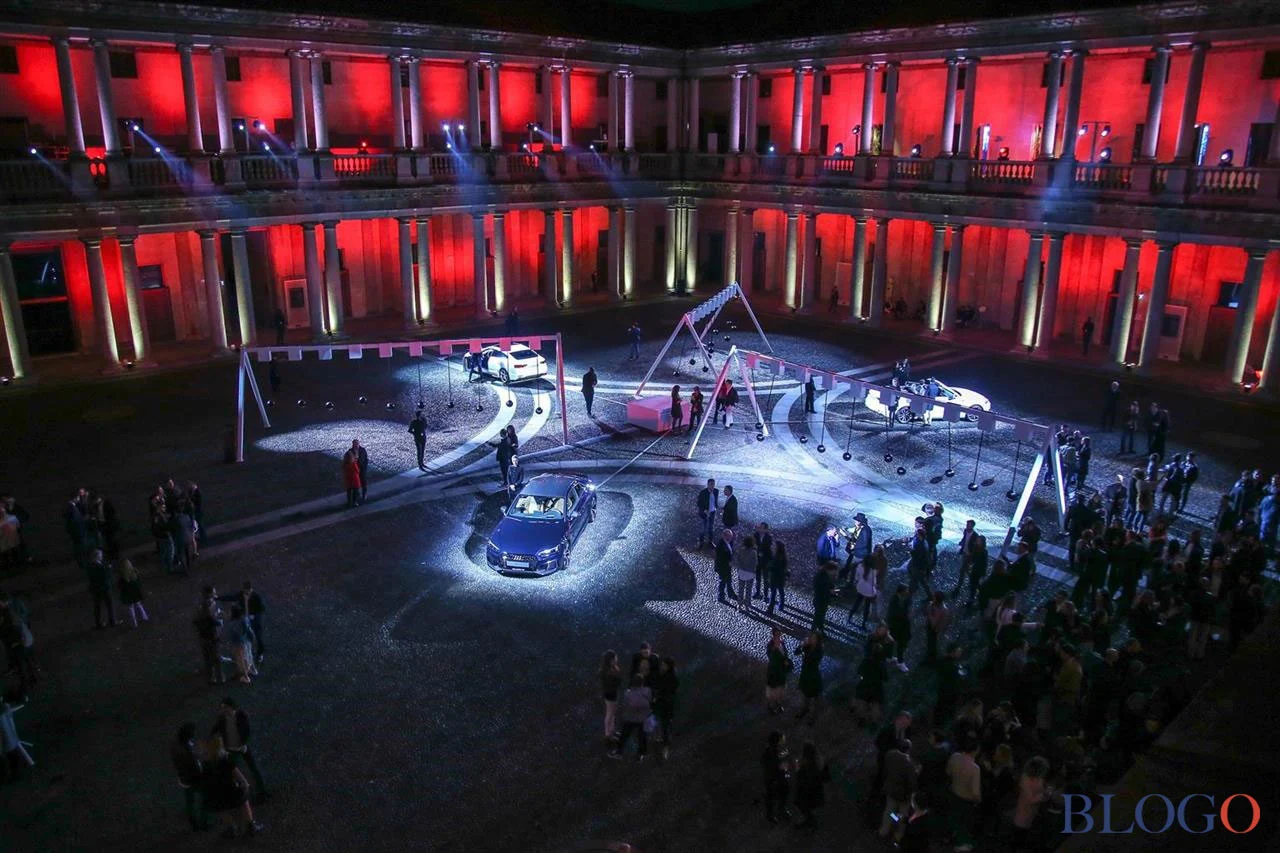 Cortile di palazzo in luce rossa con auto Audi su pedane illuminate, strutture ad altalena e visitatori di notte – Audi City Lab al Fuorisalone, Milano, Italia, 2017 – progettazione di eventi