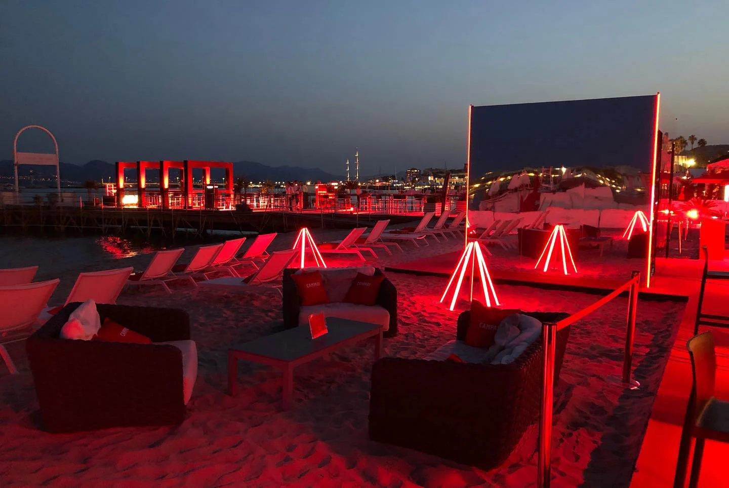 Lounge sulla spiaggia al crepuscolo con divani, treppiedi al neon rossi e un grande pannello a specchio sulla sabbia – Campari al 75° Festival di Cannes, Francia, 2022 – progettazione di eventi