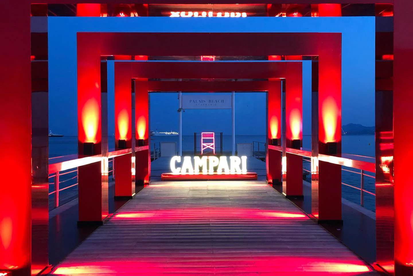 Pontile scandito da portali illuminati di rosso che conducono a un'insegna Campari luminosa sul mare al crepuscolo – Campari al 75° Festival di Cannes, Francia, 2022 – progettazione di eventi