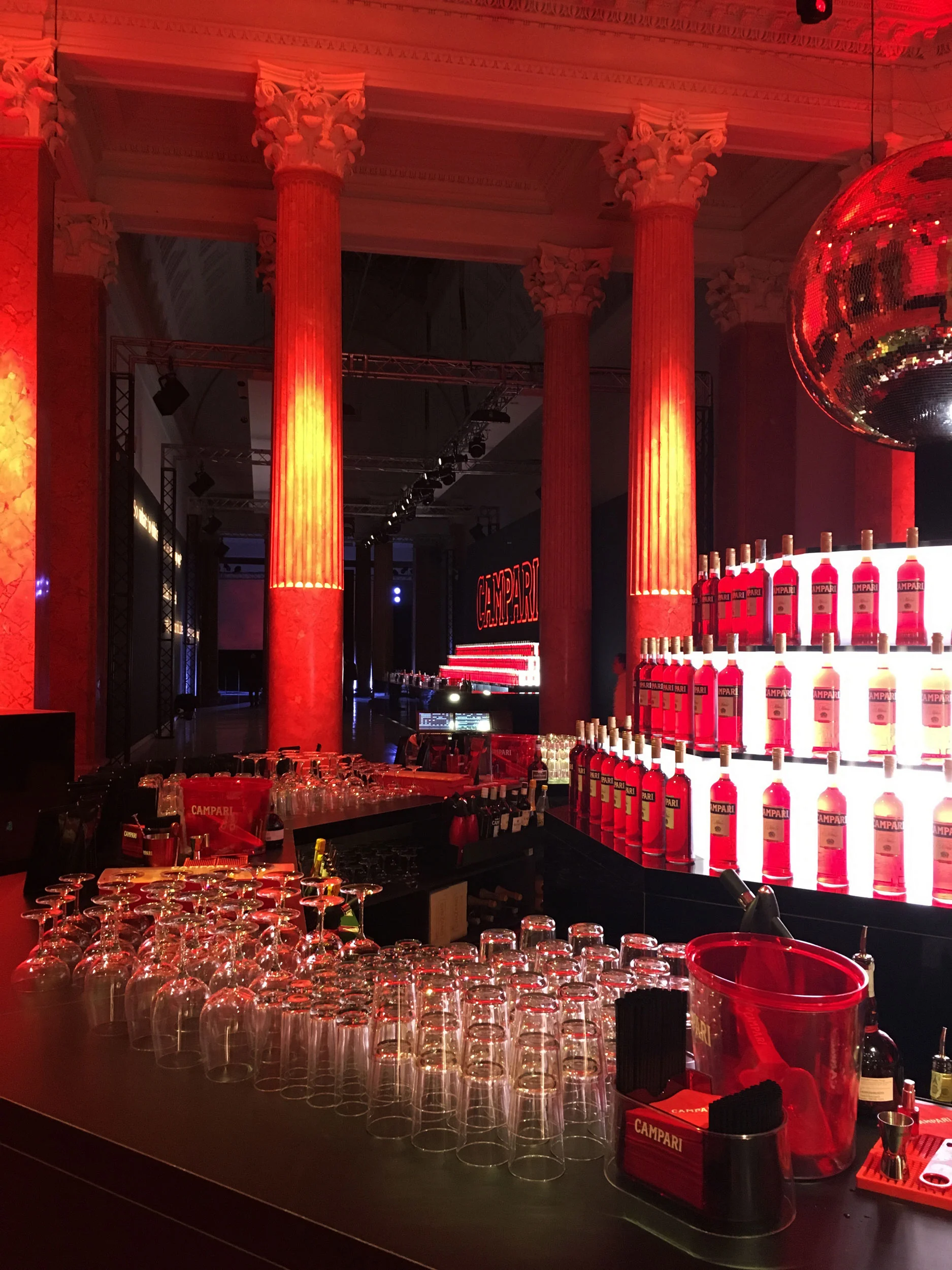 Bancone bar con file di bicchieri e bottiglie Campari retroilluminate tra colonne classiche illuminate di rosso – Campari Red Diaries, Milano, Italia, 2017 – progettazione di eventi