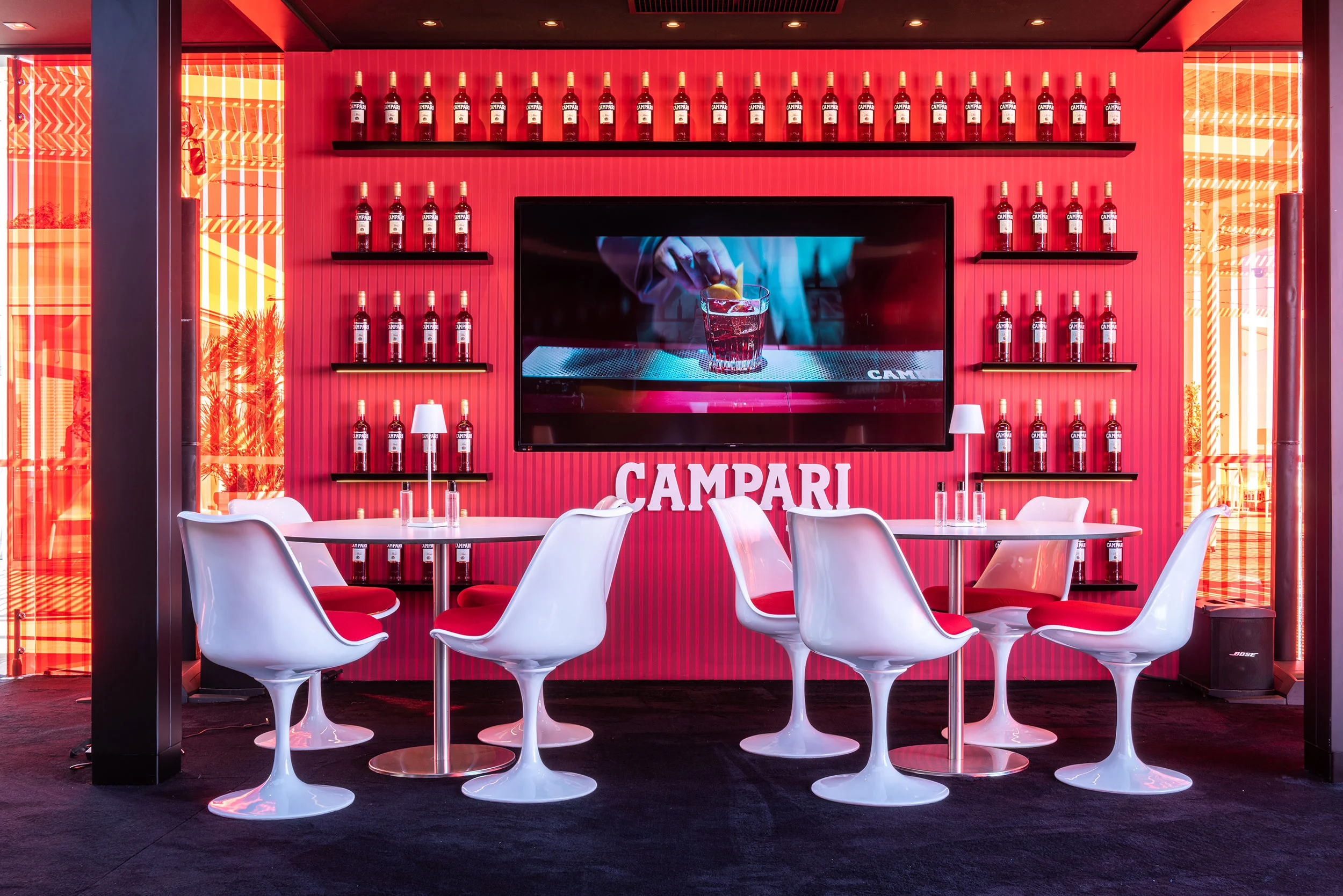Angolo lounge con sedie Tulip bianche davanti a una parete rossa di bottiglie Campari e uno schermo video – Campari Lounge Experience al Festival di Cannes, Francia, 2022 – progettazione di eventi